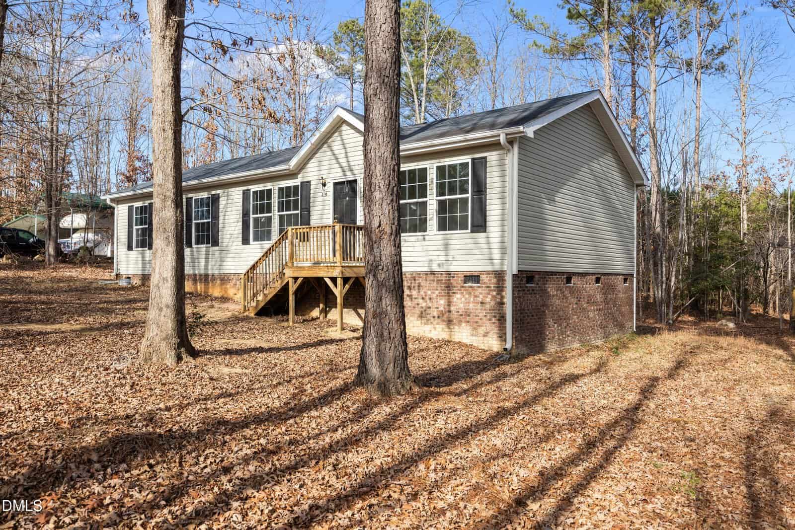 Single-story house with tan siding and brick foundation, small wooden porch with steps, surrounded by tall trees in a leaf-covered yard on a sunny day.