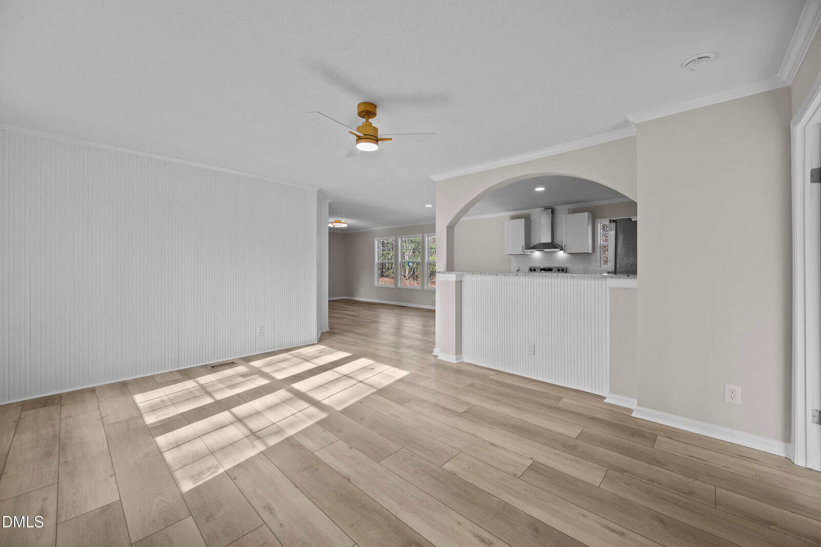 Bright, modern open living area with light wood floors, white walls, ceiling fan, arched kitchen pass-through, and large windows letting in natural light. Adjacent dining space is visible in the background.