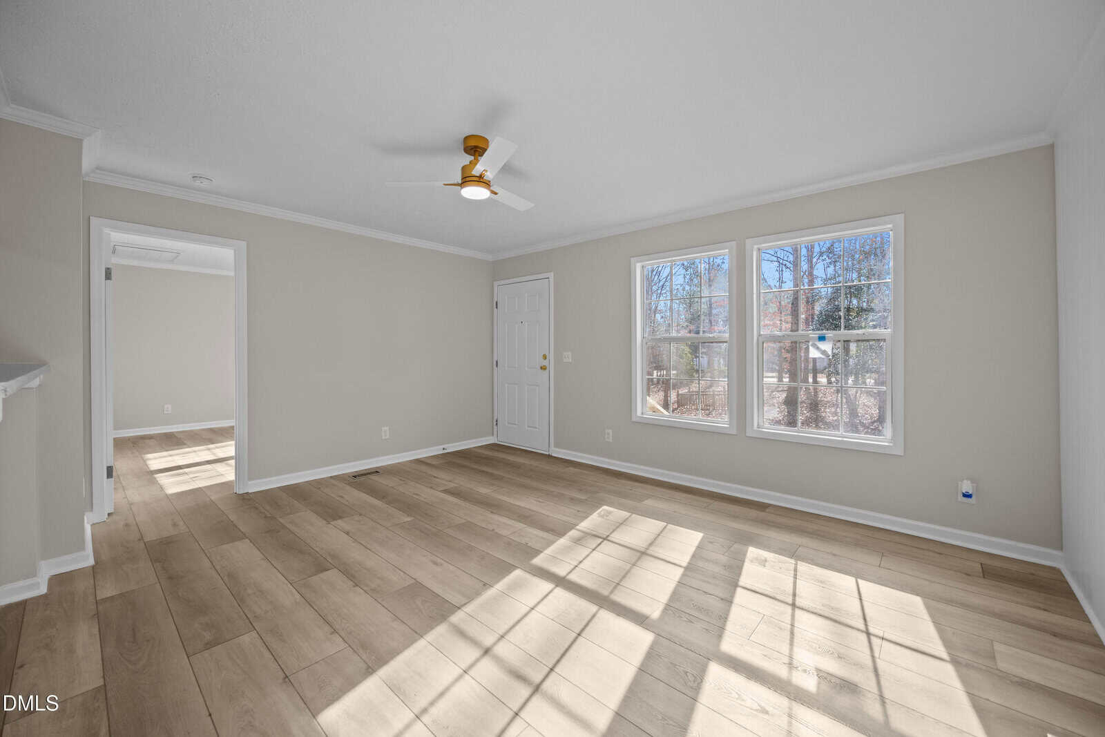 Bright, empty room with light wood floors, beige walls, two windows, white ceiling fan, and a doorway leading to another room. Sunlight streams through the windows, casting shadows on the floor.