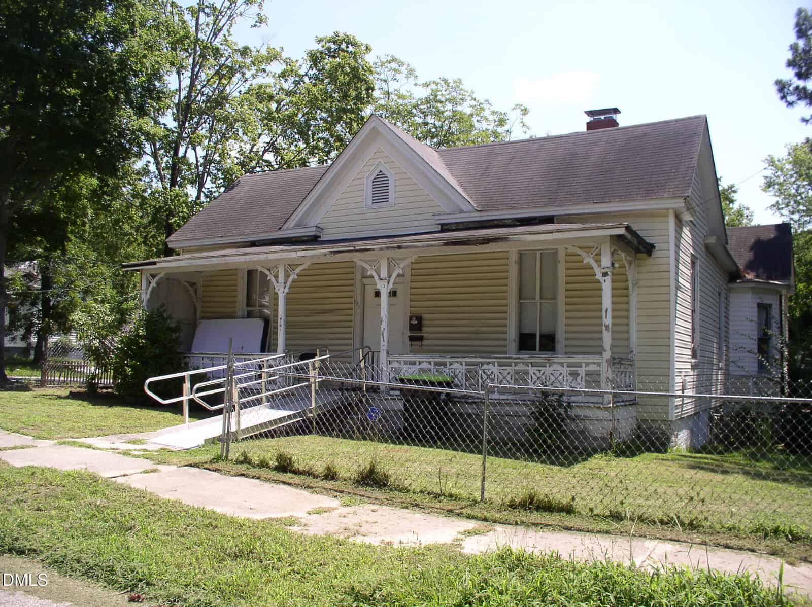 Nestled at 332 Rowland Street in Henderson, this small, single-story house features light-colored siding, a covered front porch with a wheelchair ramp, and is enclosed by a chain-link fence. Overgrown grass surrounds the home, while mature trees and bushes provide additional privacy and shade.