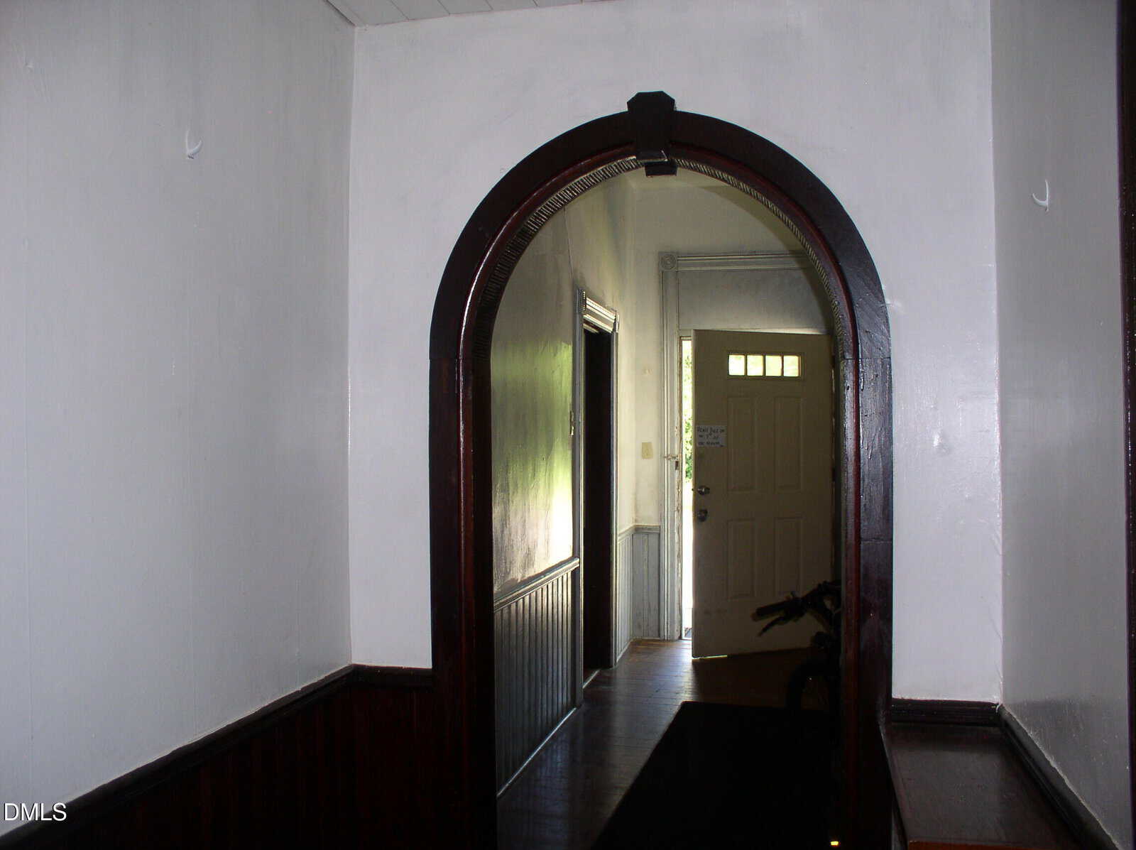 A hallway with dark wood trim and white walls features an arched doorway leading to a sunlit entryway with a partially open door. Light streams in from outside, illuminating part of the hallway floor.