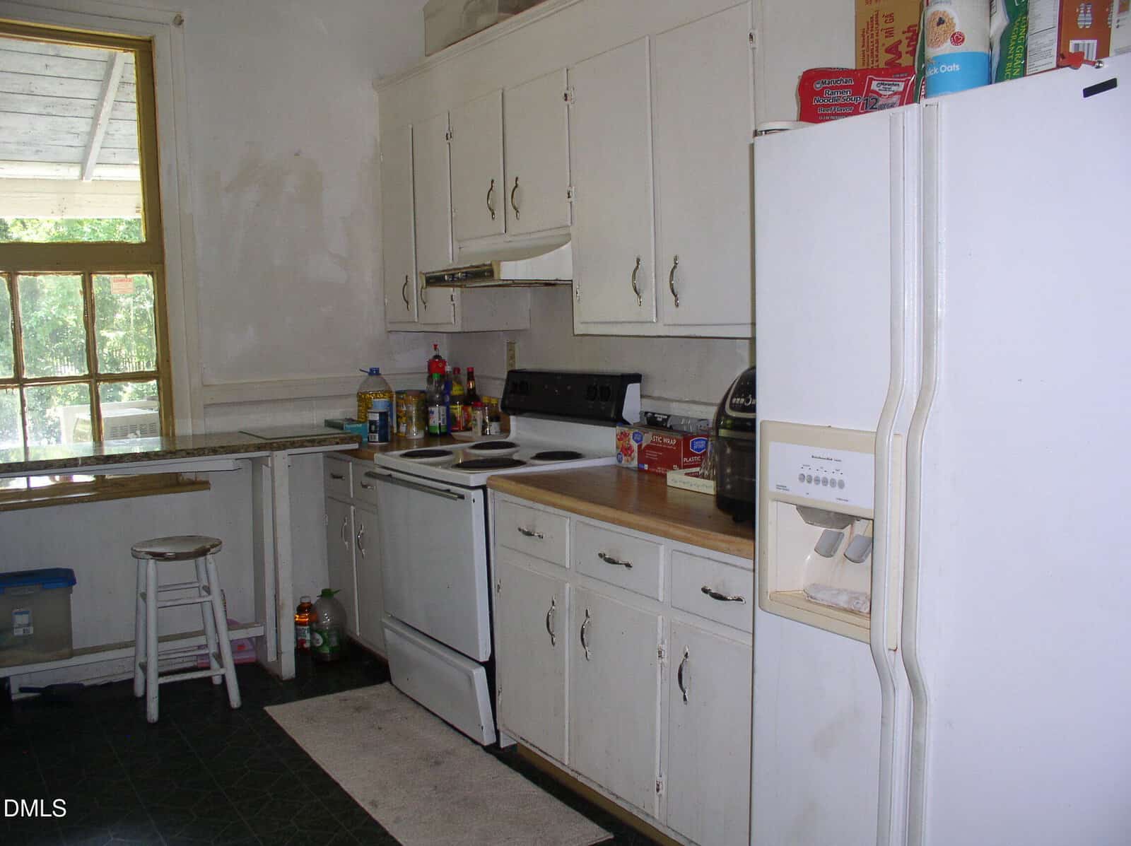 A small, older kitchen with white cabinets and appliances, a stove with items on the counter, a refrigerator, and a stool by a window. Various pantry goods are on the counter and above the cabinets.