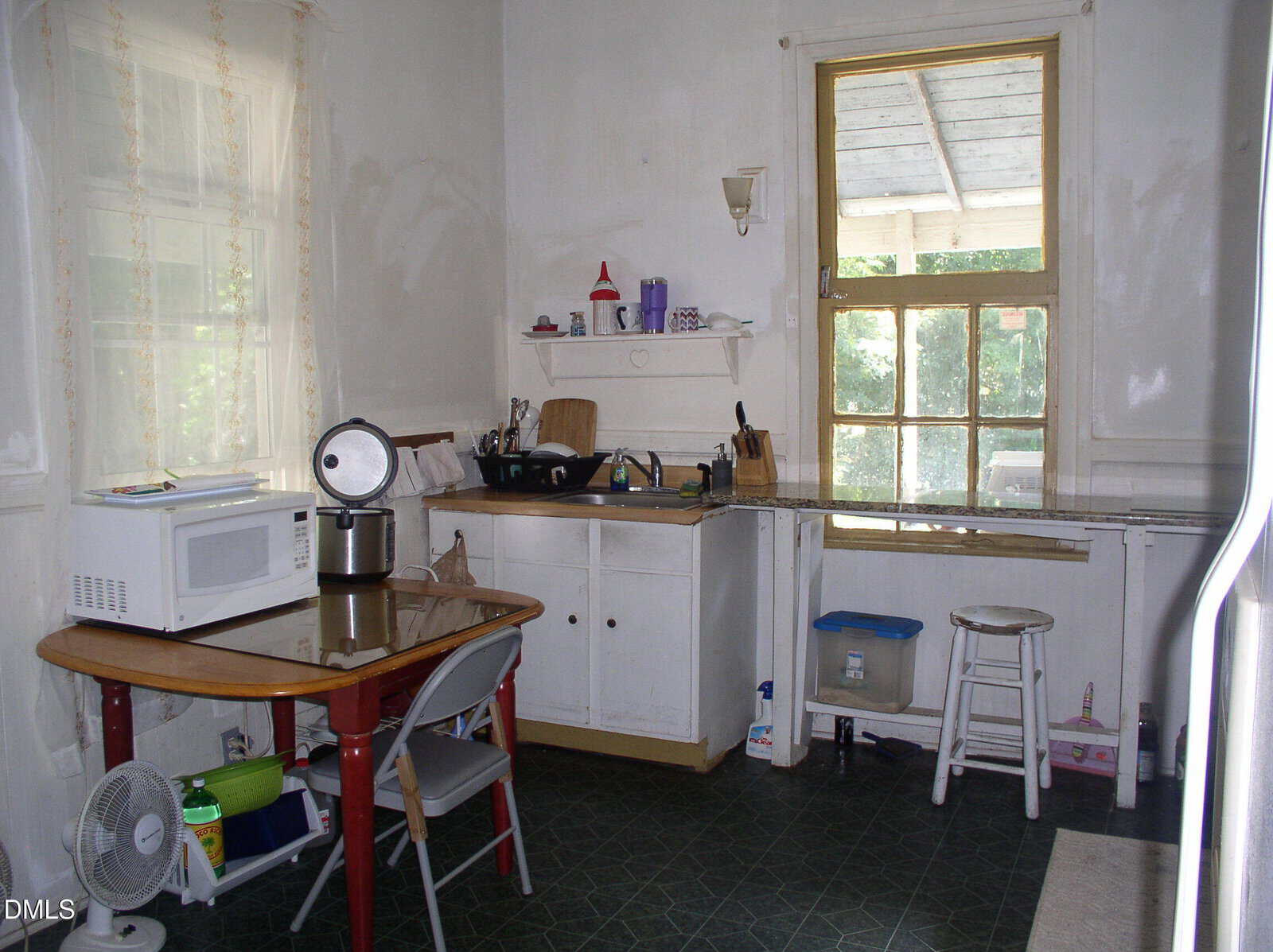 Simple kitchen with a microwave on a small table, folding chair, sink with dishes, white cabinets, and a window overlooking a porch. Various household items are on the counter and shelves. The floor is dark patterned linoleum.