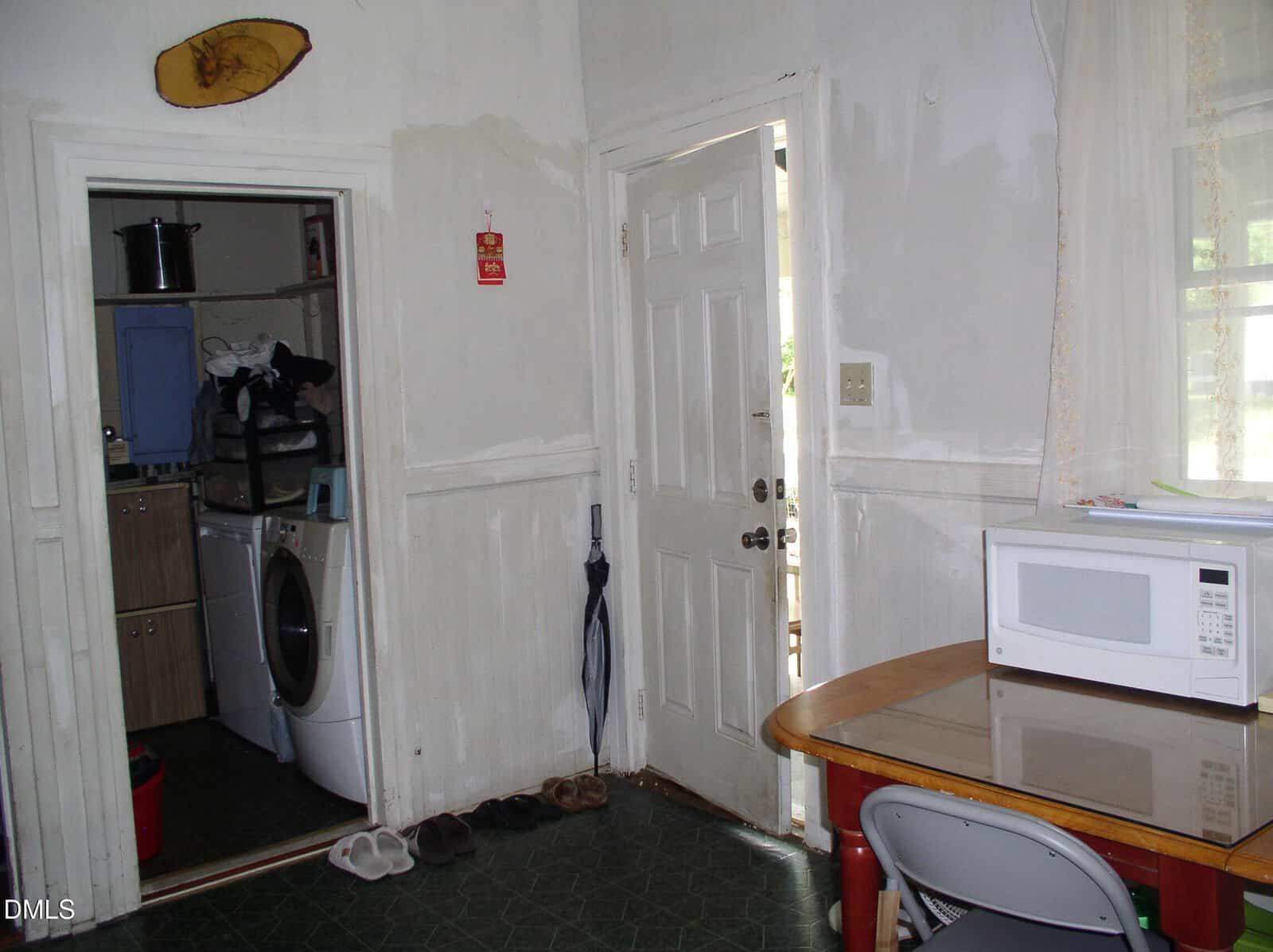 A small kitchen with a microwave on a table and a folding chair. An open door leads outside. Another door reveals a laundry area with a washing machine and shelves. Shoes and an umbrella are by the door.