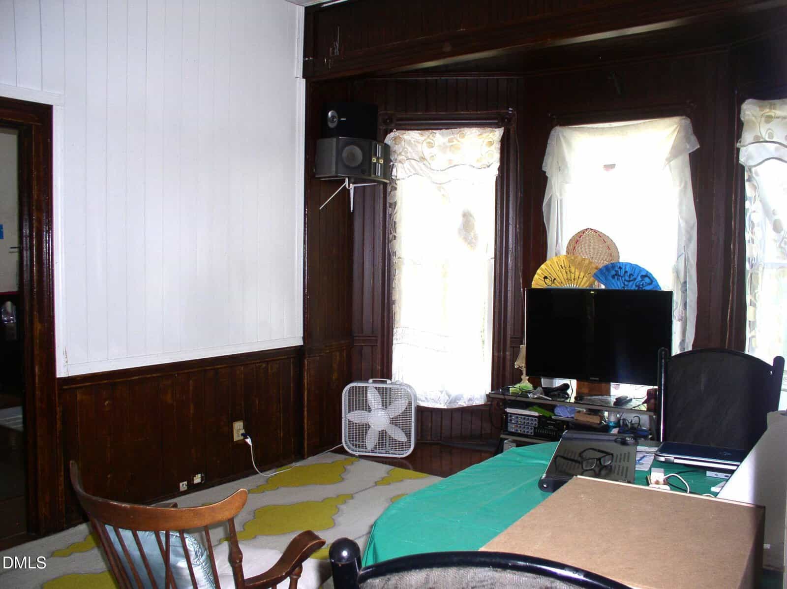 A home office with a desk, chair, computer, table fan, and window with sheer curtains. Dark wood paneling and white walls surround the room. Two decorative fans sit behind the monitor.