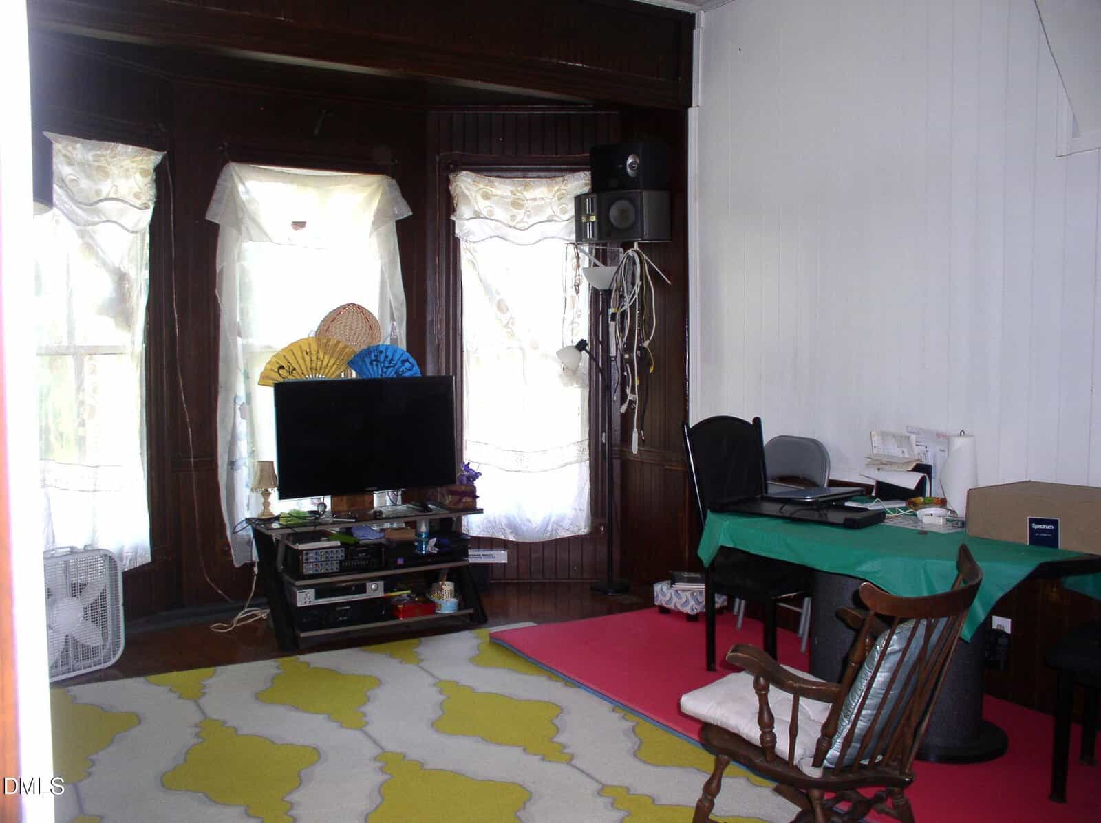 Living room with a TV, shelves, computer desk, green tablecloth, rocking chair, decorative fans, speakers, and a yellow-patterned rug. Light enters through windows with lace curtains.