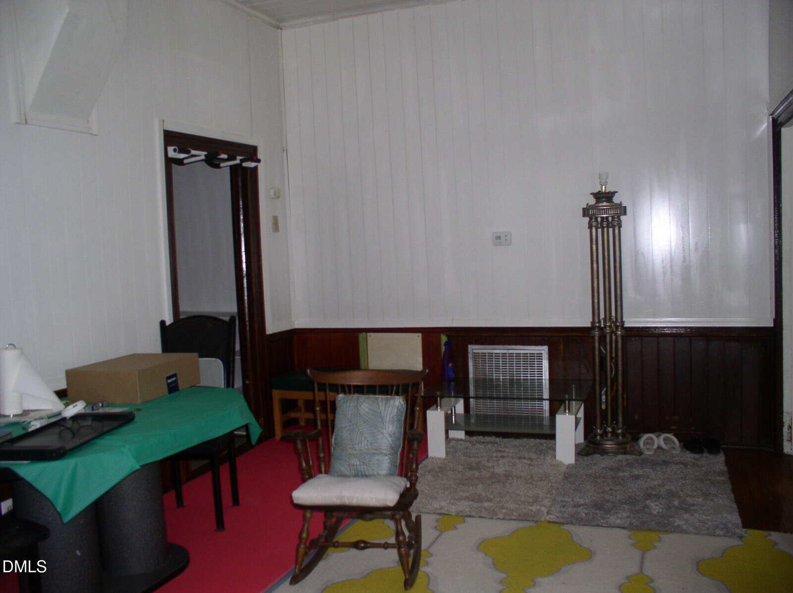 A living room with dark wood paneling, assorted chairs, a glass coffee table, a tall lamp, a heater, a cardboard box on a table, and a pink mat on the floor.