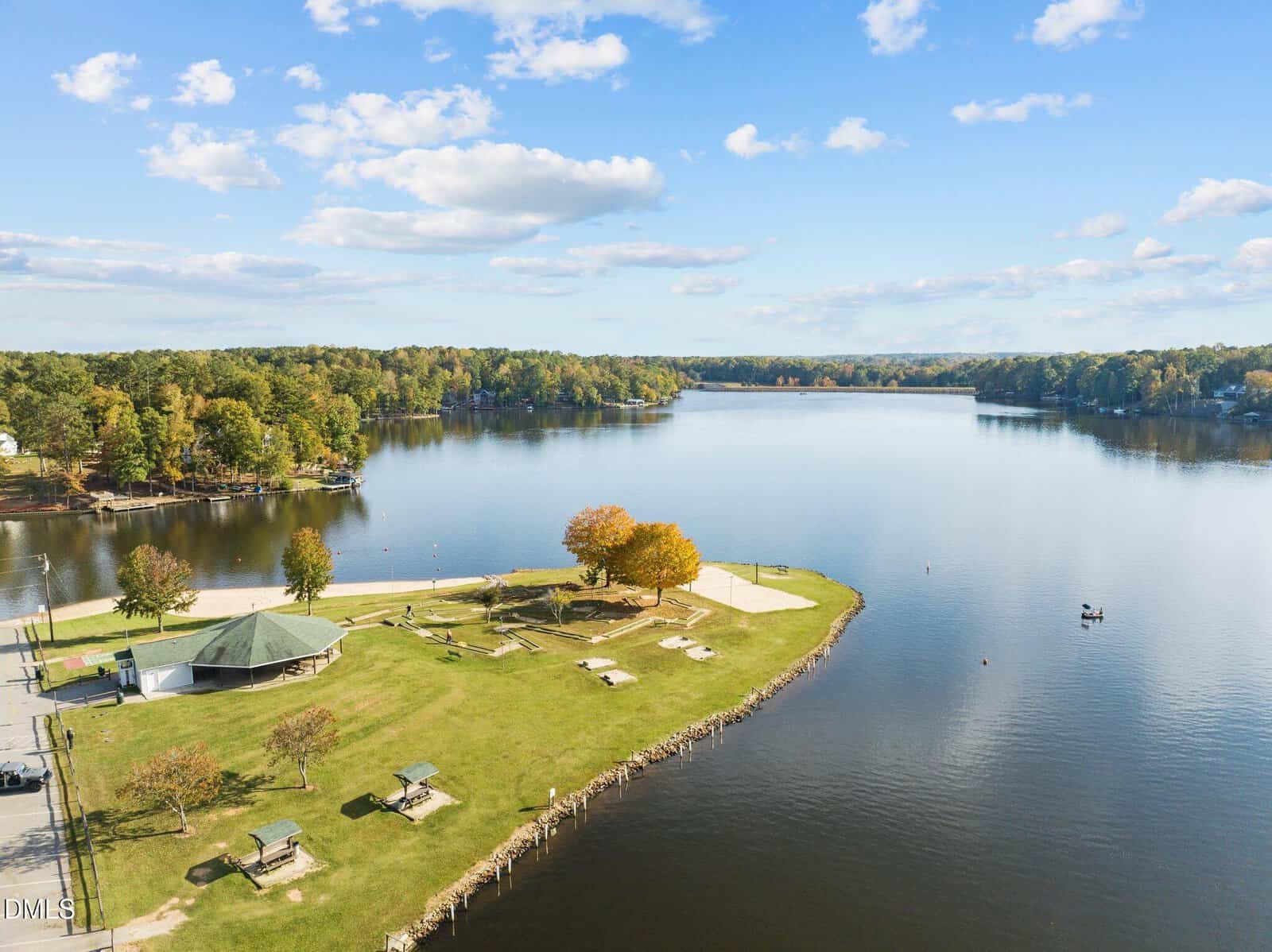 Aerial view of a lakeside park with picnic tables, a pavilion, trees with autumn foliage, and a small beach. Calm lake waters reflect a blue sky with scattered clouds, surrounded by forested shoreline.