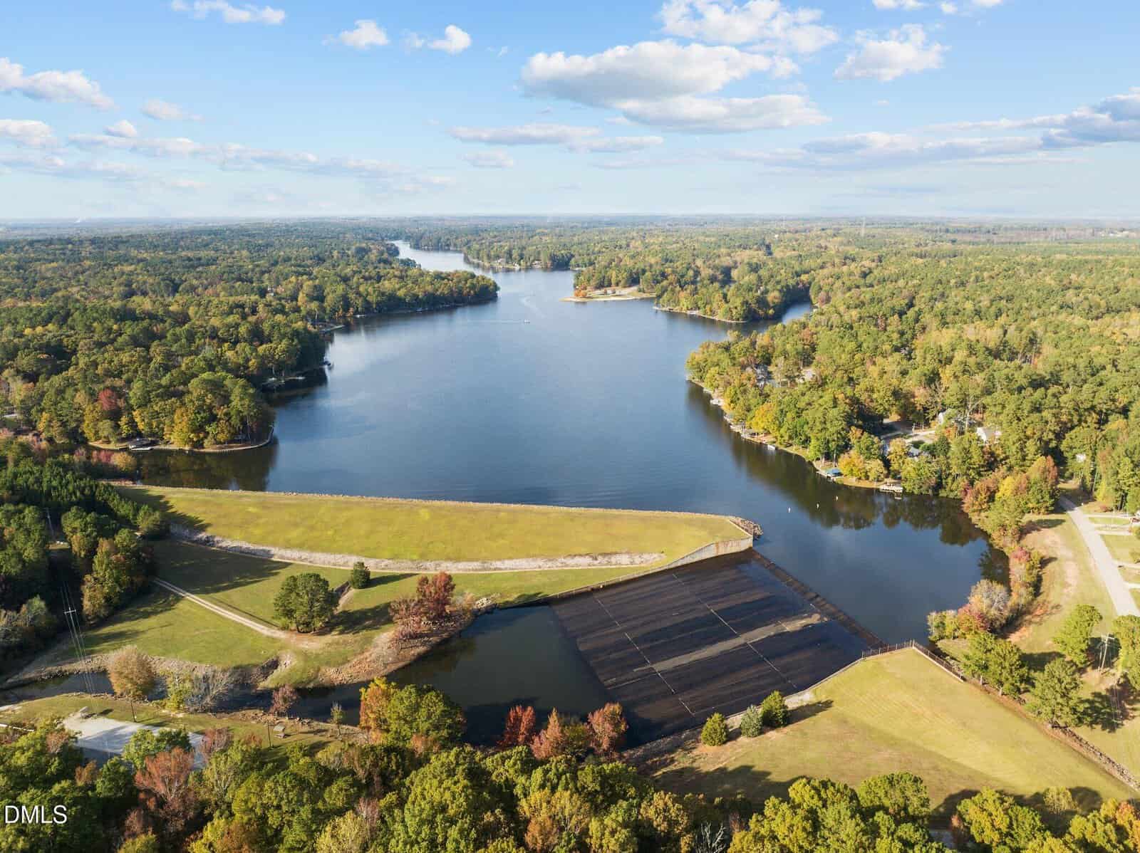Aerial view of a large, calm lake surrounded by dense green forest, with a dam and open fields in the foreground under a blue sky with scattered clouds.