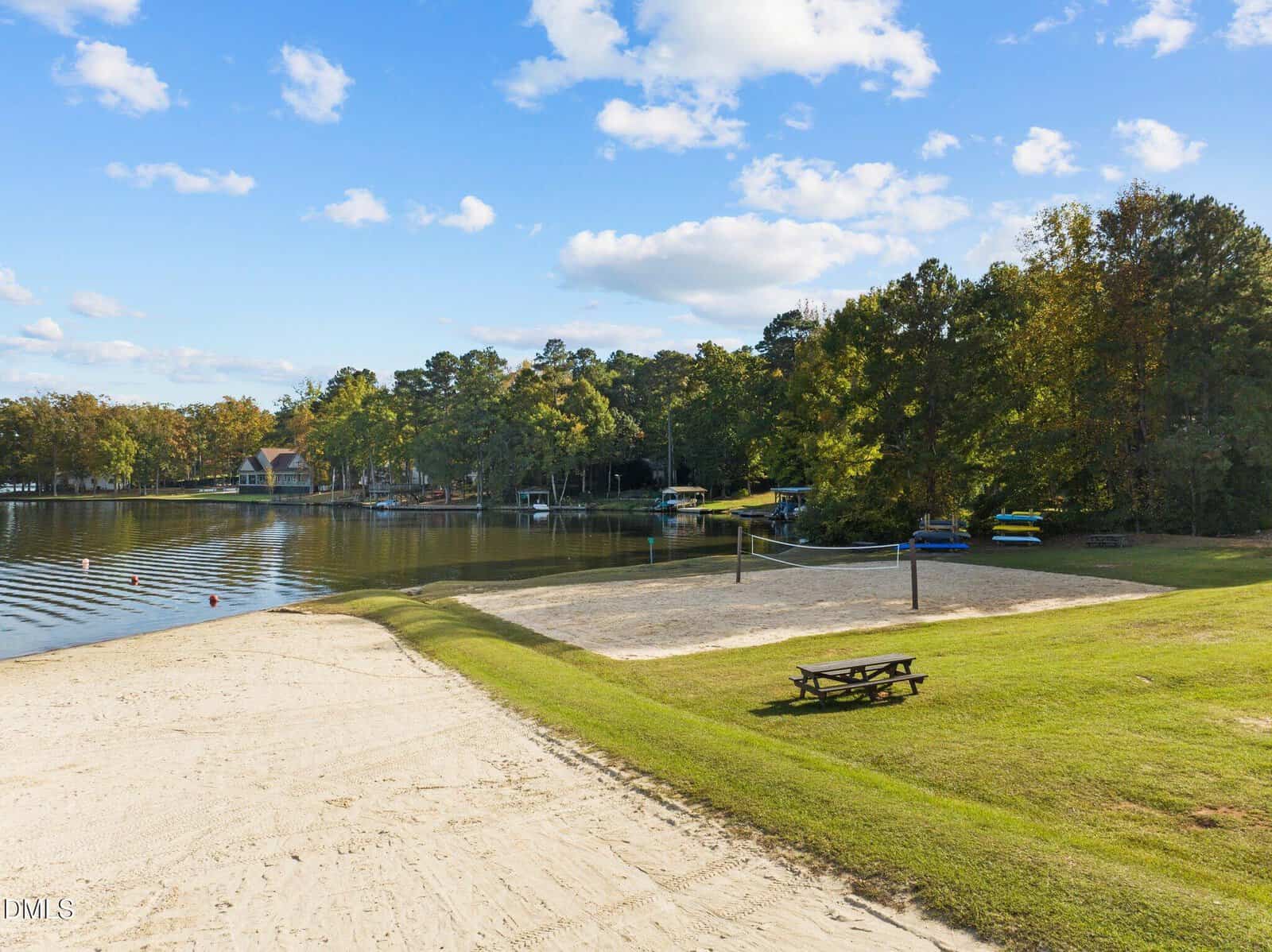 A sandy beach with a volleyball court, picnic table, calm lake, trees, docks, and boats under a blue sky with scattered clouds.