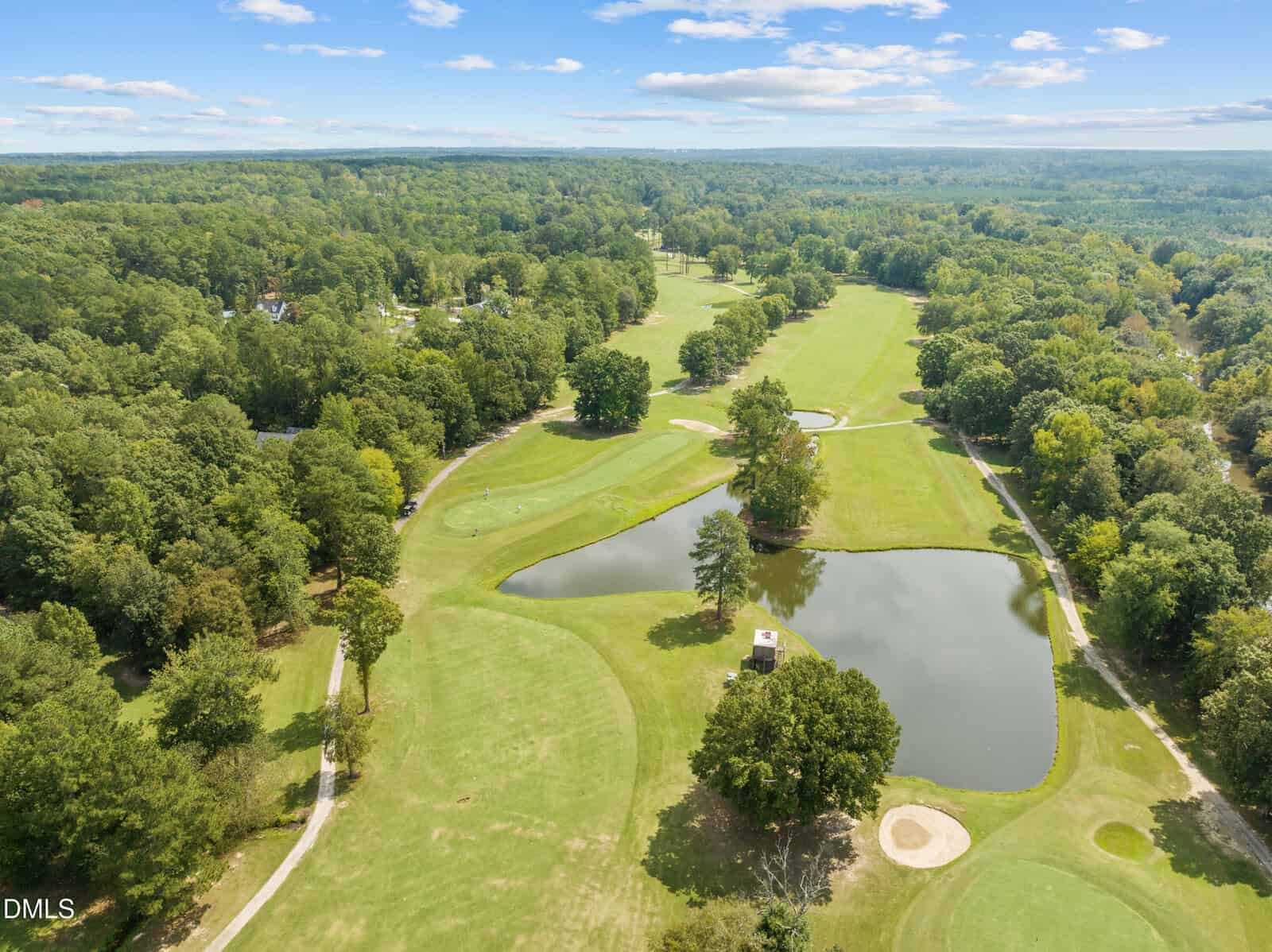 Aerial view of a golf course with green fairways, sand bunkers, a small pond, clusters of trees, and surrounding forest under a partly cloudy sky. Golf cart paths wind through the landscape.