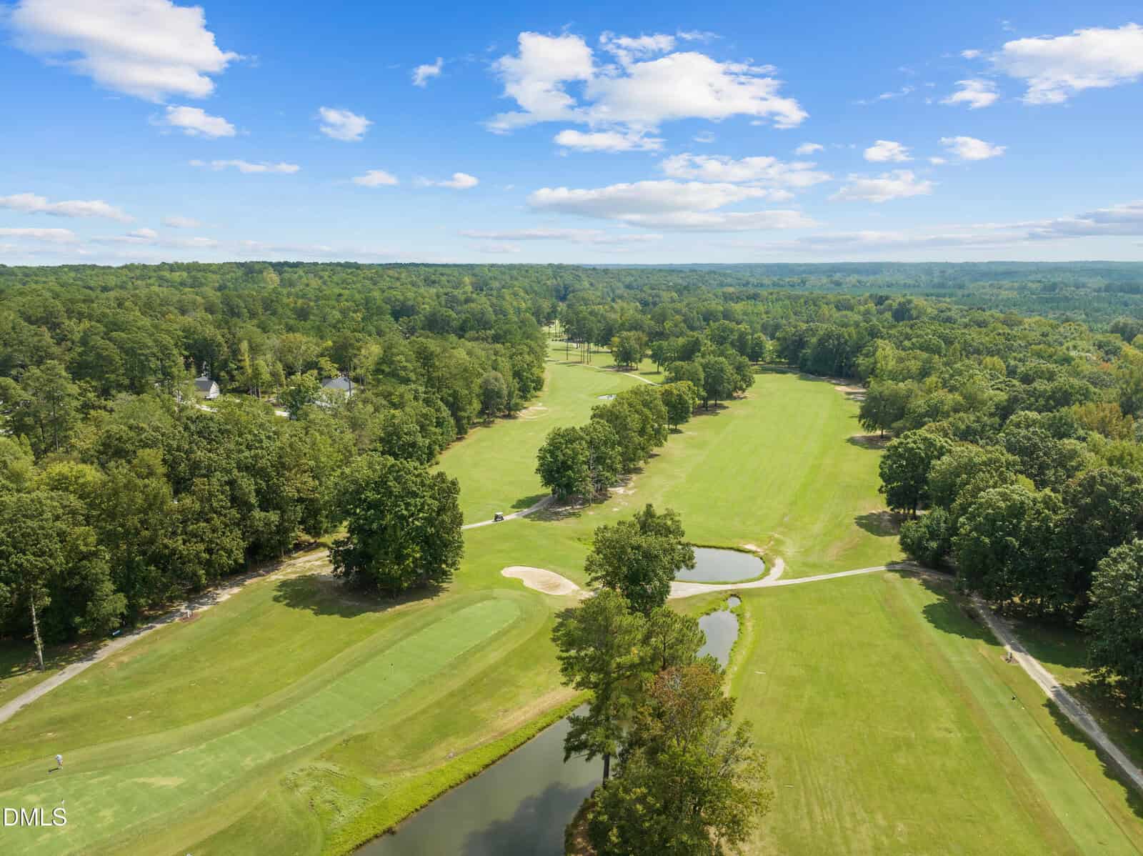 Aerial view of a golf course with green fairways, putting greens, sand bunkers, small ponds, and surrounding trees under a blue sky with scattered clouds.