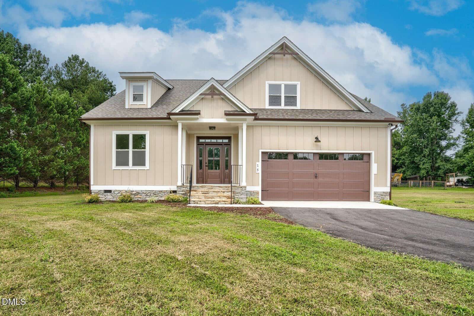 A modern two-story house at 15 Black Feather Lane, Youngsville, featuring beige siding, white trim, a front porch with stone steps, and a double garage. The lawn is mowed, and trees line the background under a partly cloudy sky.