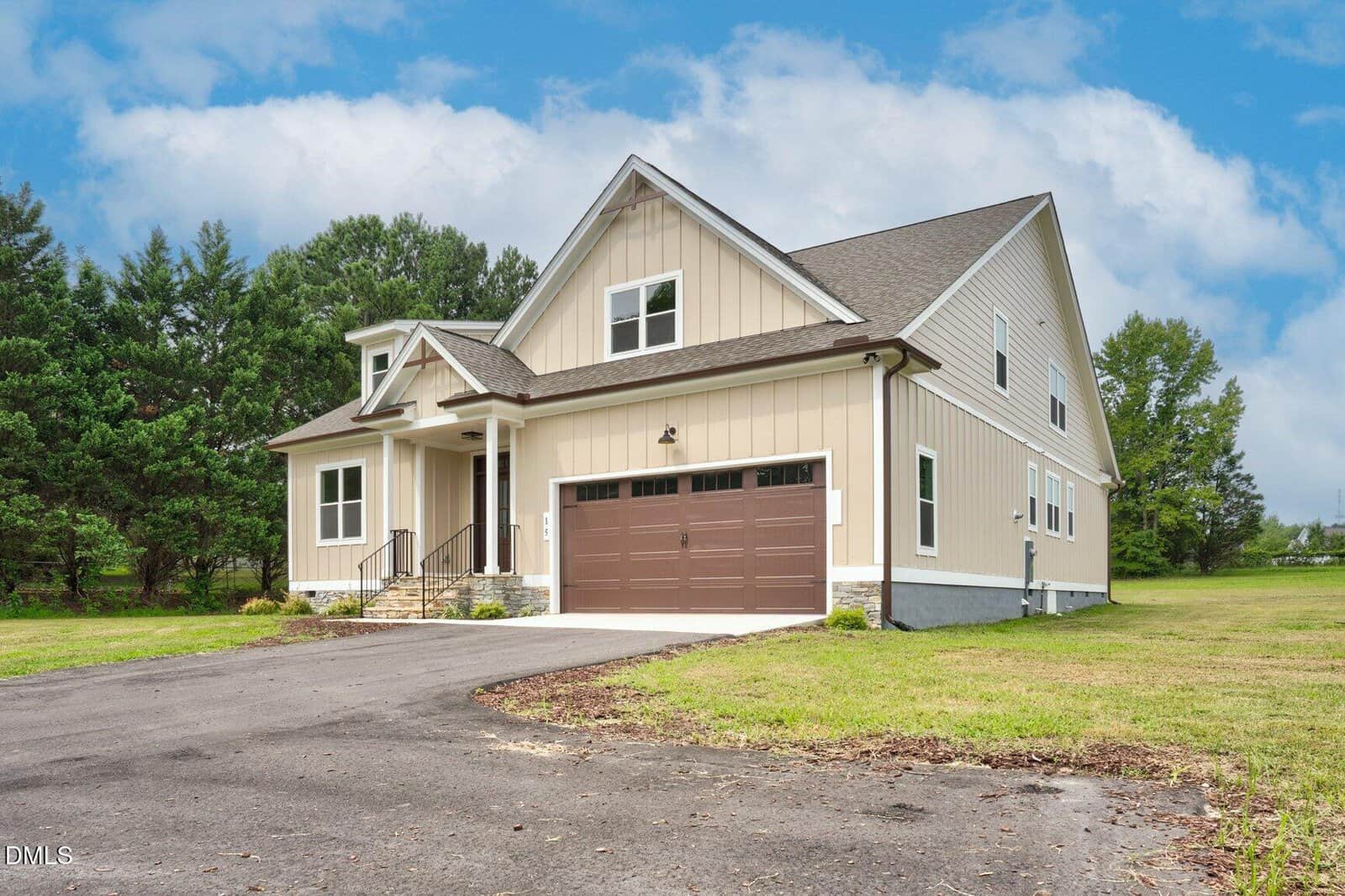 A modern two-story house with beige siding, a brown double garage door, small front porch, and a paved driveway, surrounded by grass, trees, and a partly cloudy sky overhead.