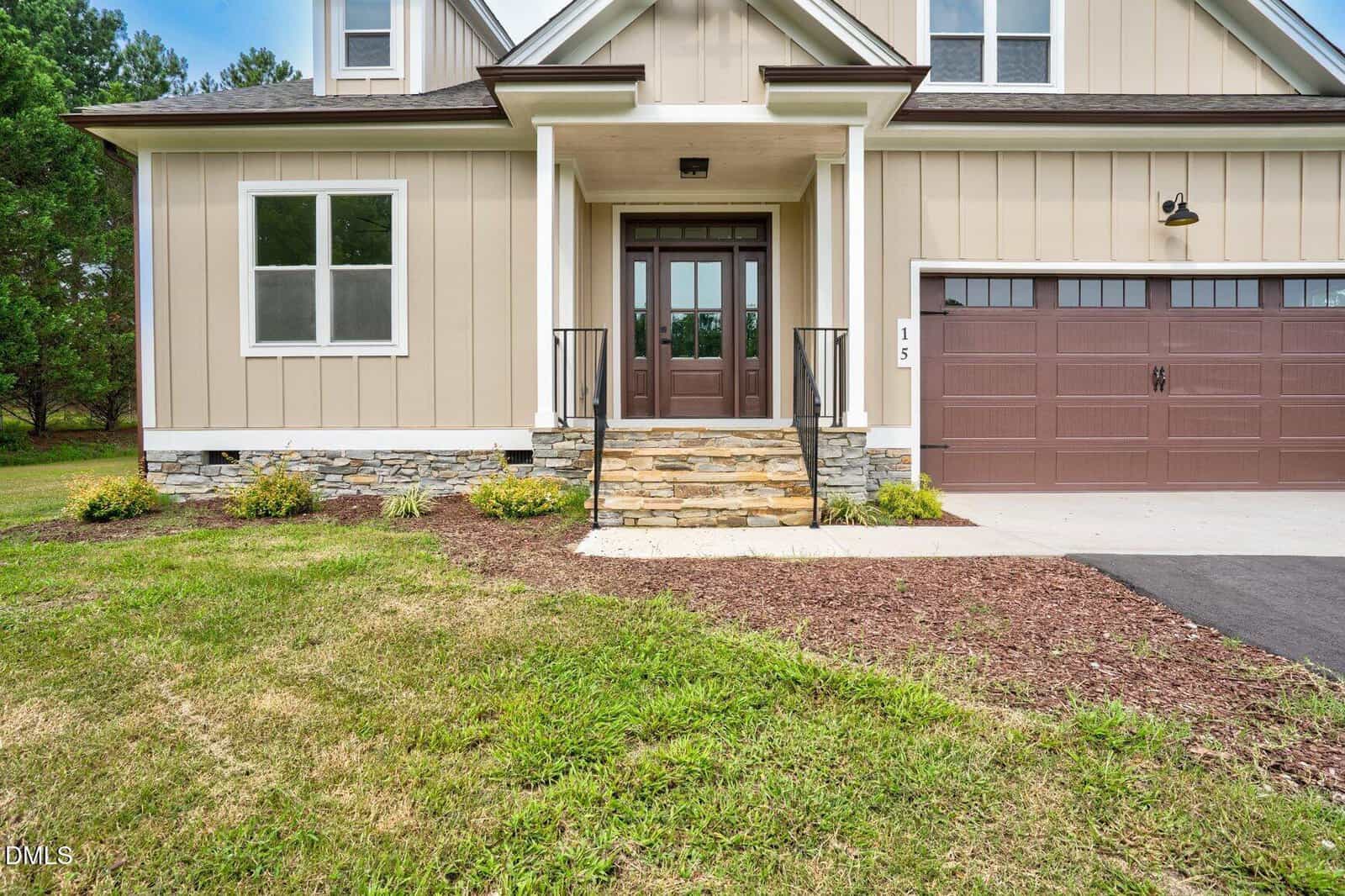 Front view of a beige house with stone accents, a covered front porch with steps, double front doors with glass panels, and an attached brown garage door. The yard has grass and a mulch bed with small plants.