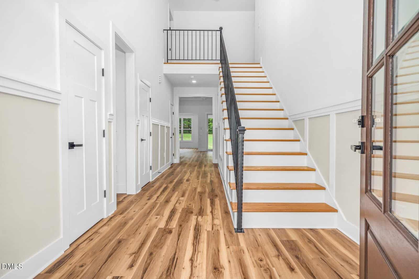 A bright entryway with light wood floors, white walls, a staircase with black railing, and doors leading to other rooms. Natural light comes from windows at the end of the hall.