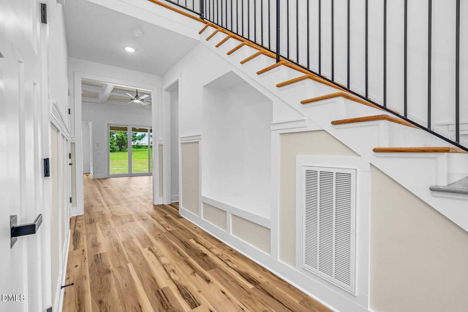 A hallway with light wood flooring, white walls, built-in shelves under a staircase with a black railing, and a view into a bright living room with glass doors opening to a green lawn.