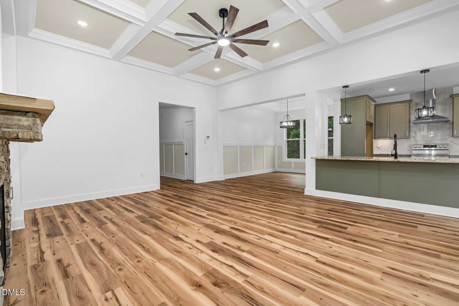 Spacious modern living area with light wood floors, coffered ceiling, ceiling fan, open kitchen with pendant lights, and dining space; white walls and stone fireplace.