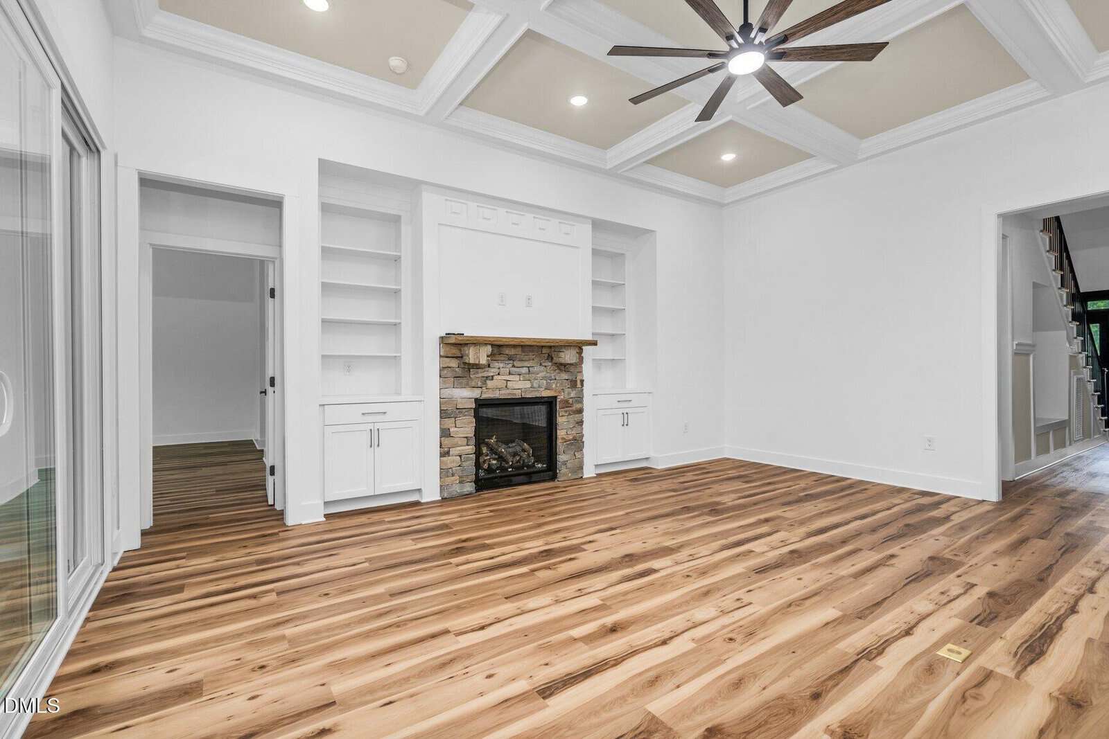 Spacious living room with hardwood floors, coffered ceiling, ceiling fan, stone fireplace, built-in white shelving and cabinets, and sliding glass doors.