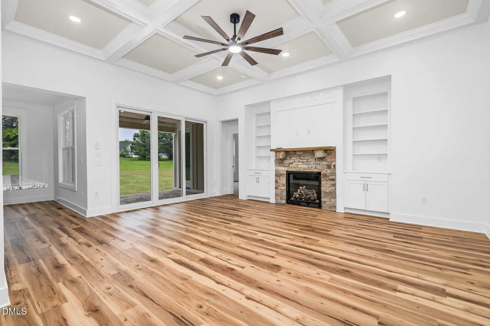 Bright living room with wood floors, white walls, coffered ceiling, ceiling fan, built-in shelves, stone fireplace, and large glass doors leading to a grassy backyard.