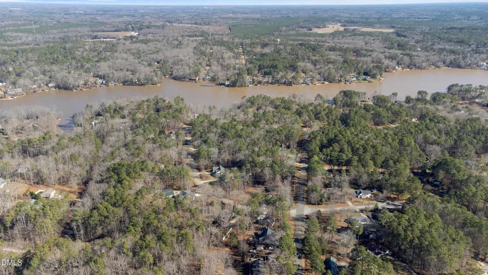 Aerial view of a wooded residential area beside a wide, muddy river or lake near Louisburg, with scattered houses and tree-lined roads—including 124 Shoshone Drive—surrounded by forested land stretching into the distance.