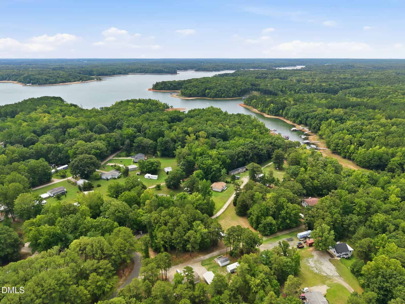 Aerial view of a lake surrounded by dense green forest, with scattered houses and roads in a rural residential area. The shoreline curves along several coves and inlets under a blue sky with light clouds.