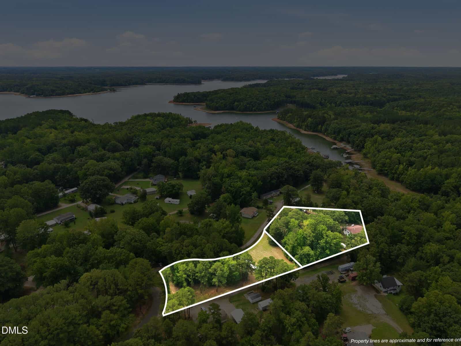 Aerial view of a wooded lakeside neighborhood with a property outlined in white, showing trees, open clearings, and nearby homes. A large lake with wooded shores is visible in the background.