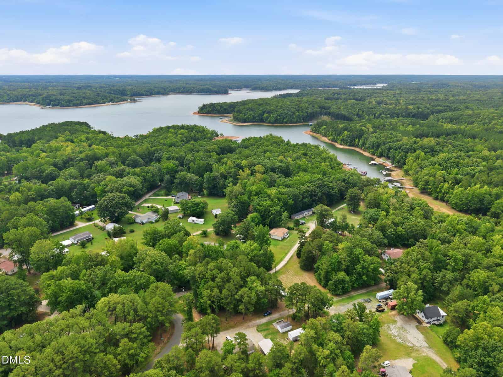 Aerial view of a lakeside community surrounded by dense green trees, houses scattered among the foliage, with a large lake and forested shoreline visible under a partly cloudy sky.