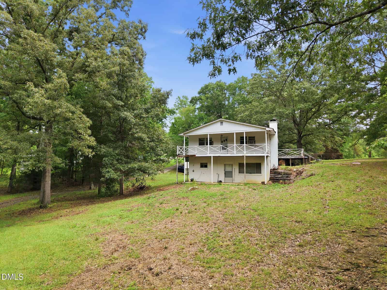 A white two-story house with a wraparound porch sits on a grassy, sloped lot surrounded by mature trees under a clear sky. Dirt patches are visible in the yard, and a driveway curves through the trees on the left.