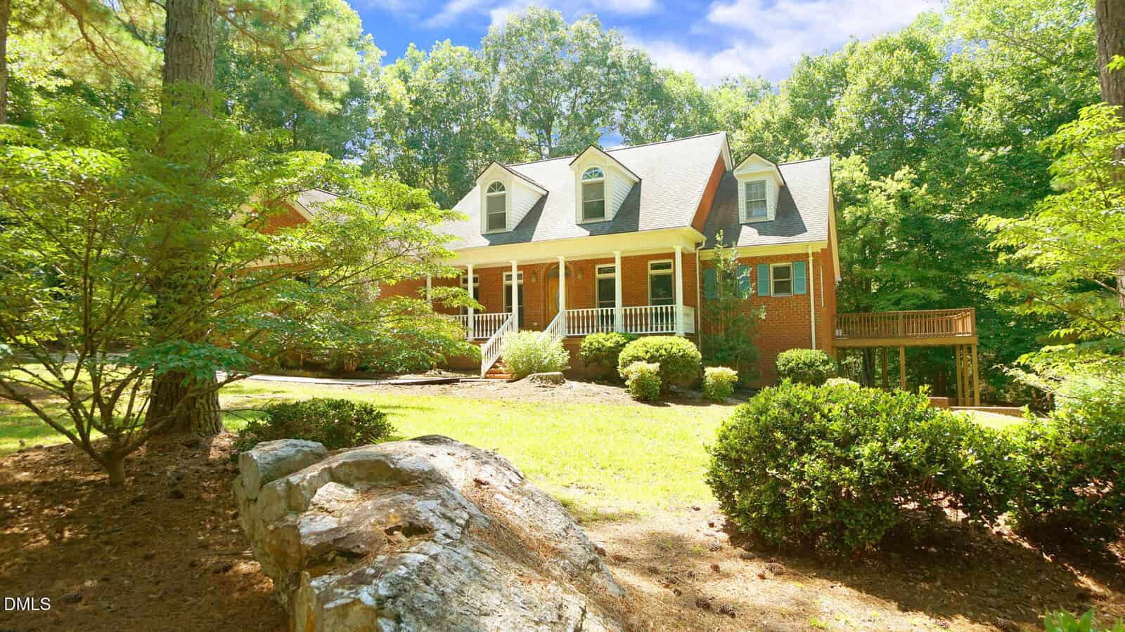Two-story brick house with white trim and dormer windows, featuring a front porch with columns, surrounded by green trees, bushes, and large rocks on a sunny day.