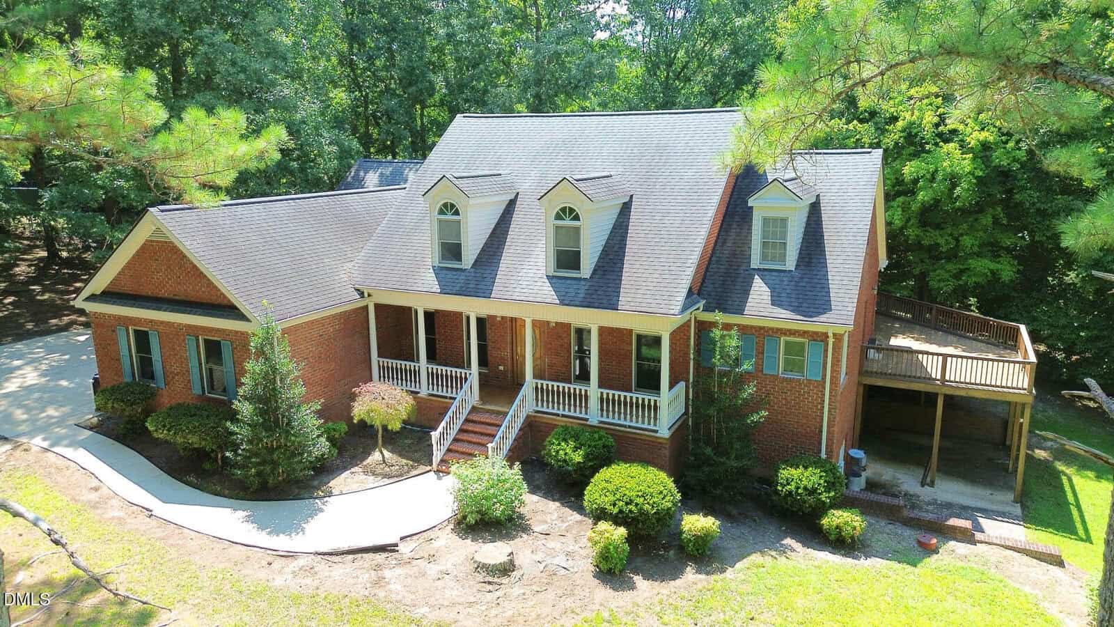 Two-story brick house with dormer windows, a covered front porch, white trim, and a side deck. Located at 1420 NC 98 Highway E in Spring Hope, the home is surrounded by greenery, shrubs, and trees, with a curved concrete walkway leading to the entrance.