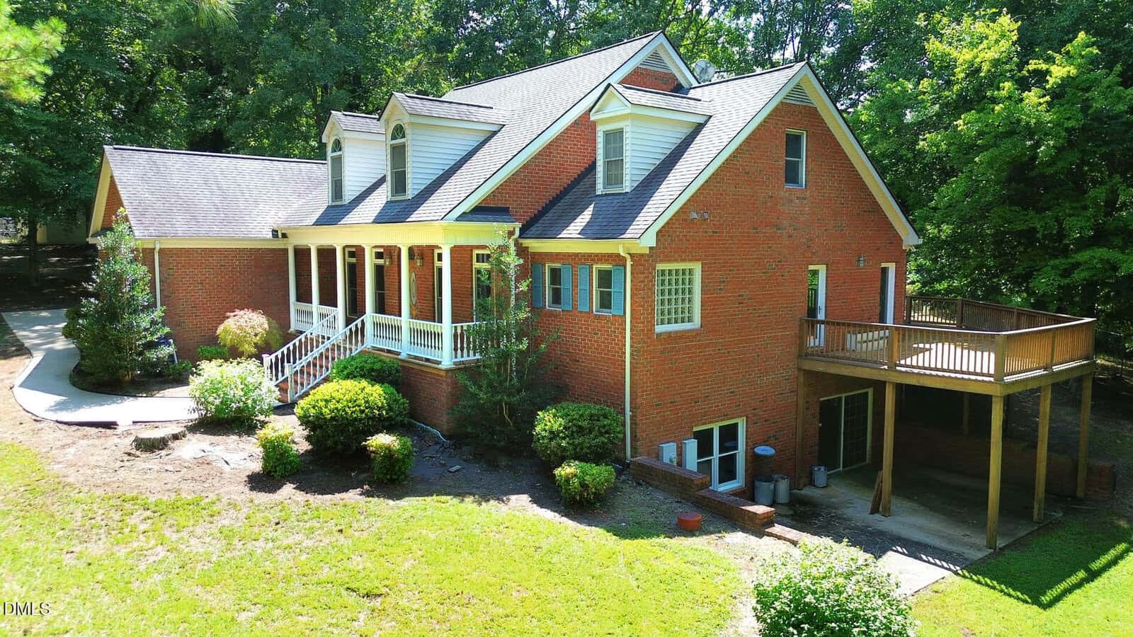 A two-story red brick house with white trim, a covered front porch, and an elevated wooden deck. The yard is landscaped with bushes and small trees, and there is a wooded area in the background.