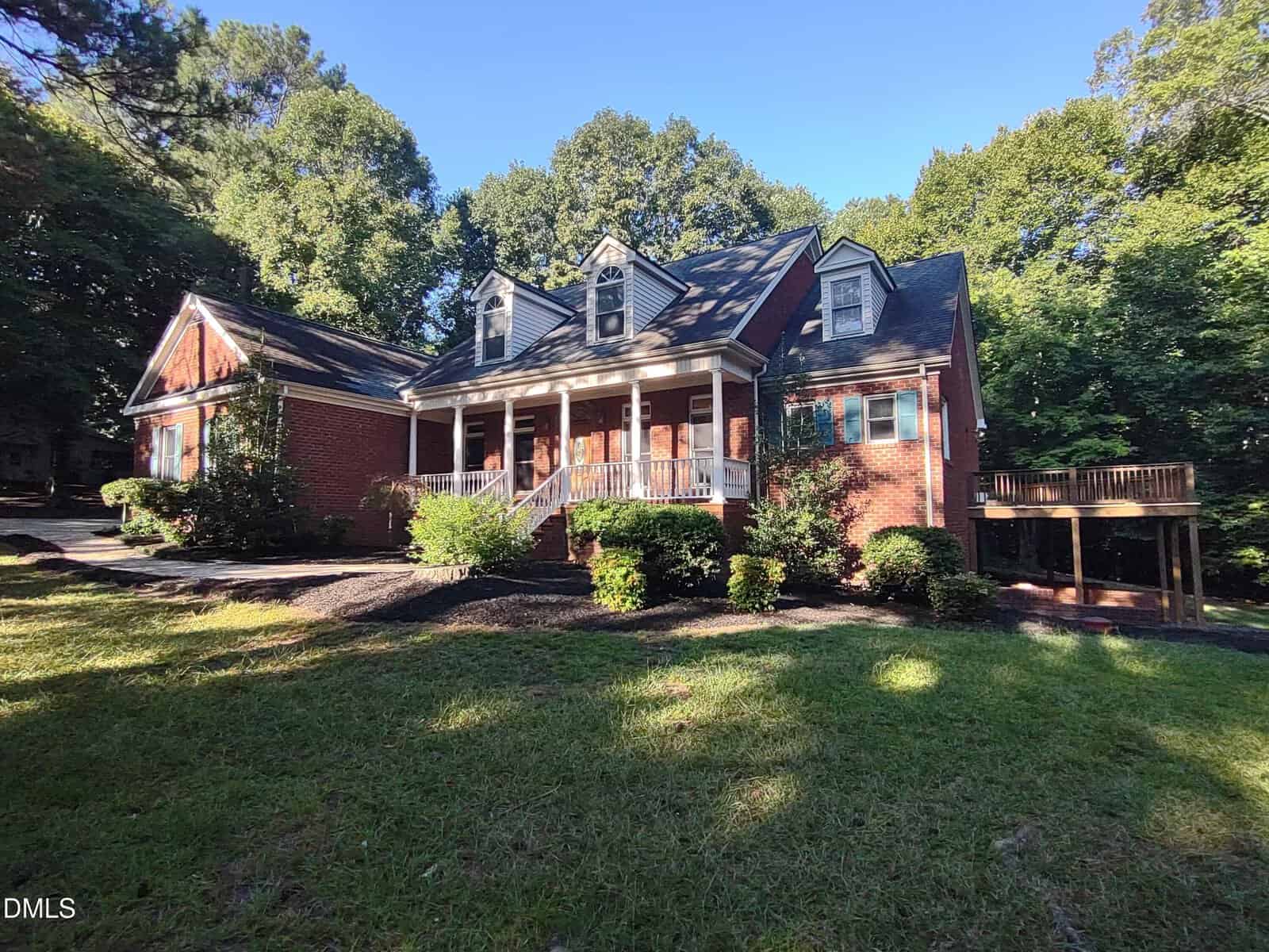A red brick house with a large front porch, dormer windows, and attached garage, surrounded by trees and greenery under a clear blue sky, welcomes you at 1420 Nc-98 in Spring Hope.