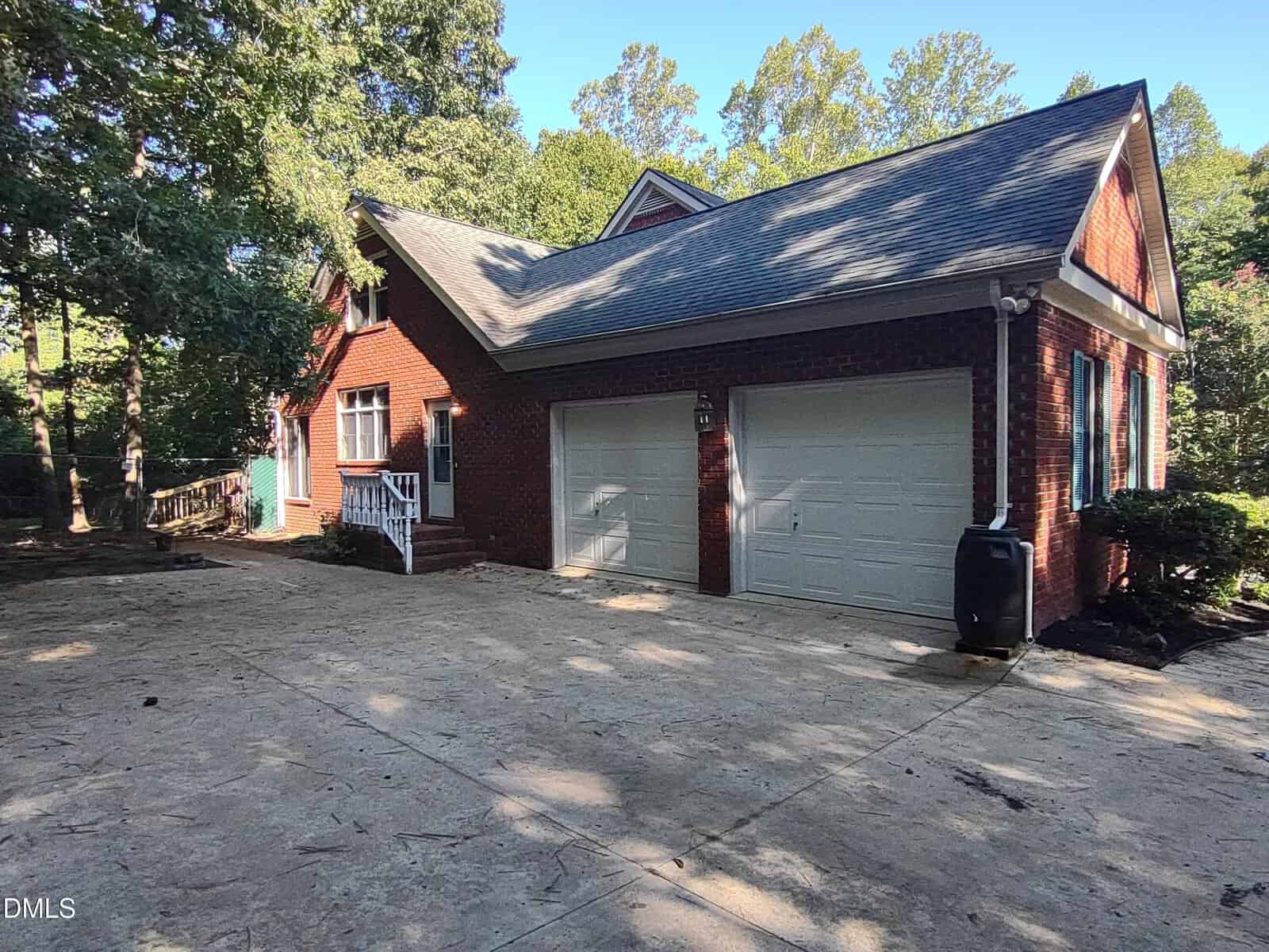 A red brick house with white trim, double garage doors, a sloped roof, and a wide concrete driveway, surrounded by trees on a sunny day.