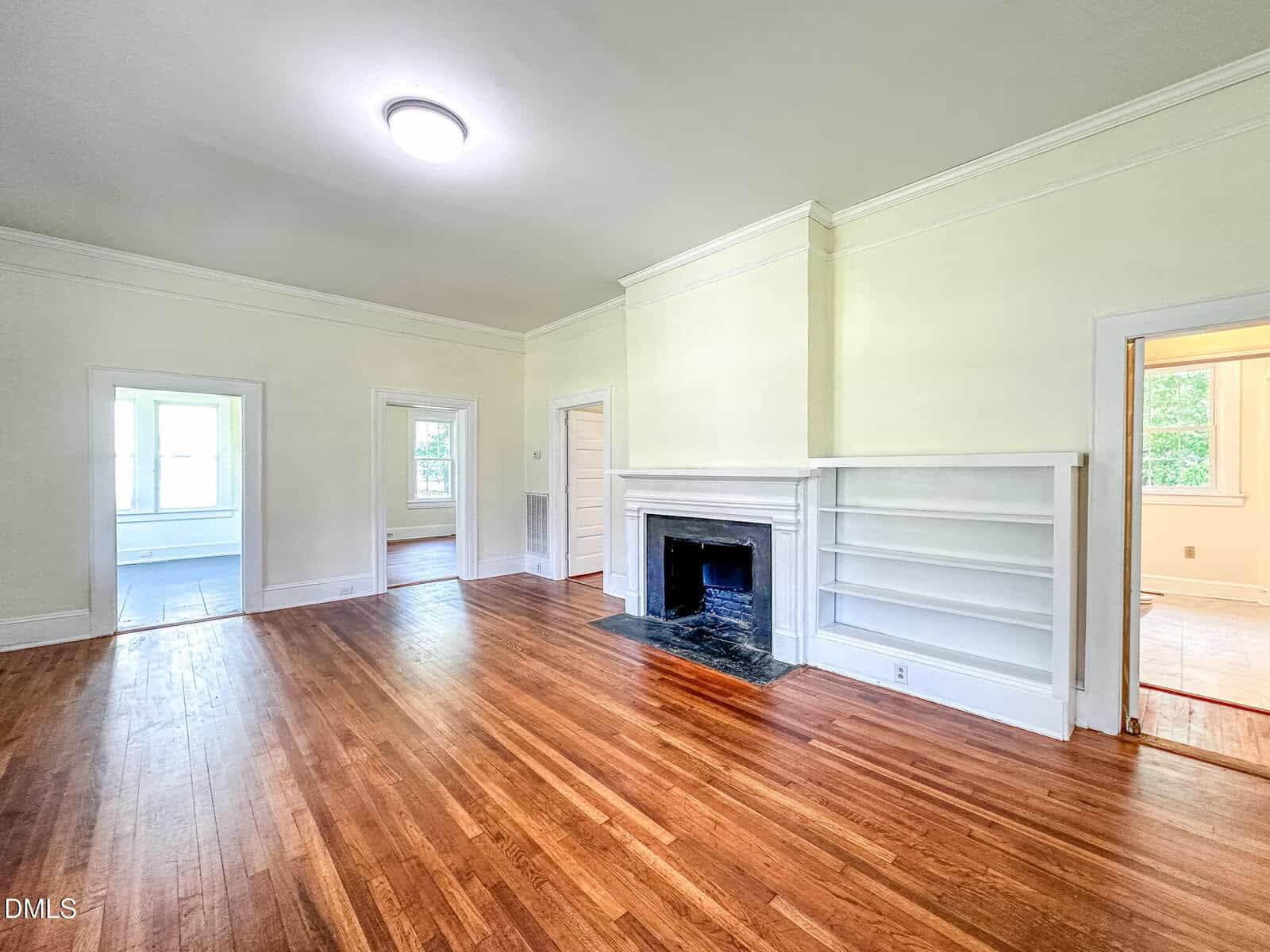 Bright living room with hardwood floors, a white fireplace with built-in shelves, cream-colored walls, and multiple doorways leading to other rooms. Ceiling light and several windows let in natural light.