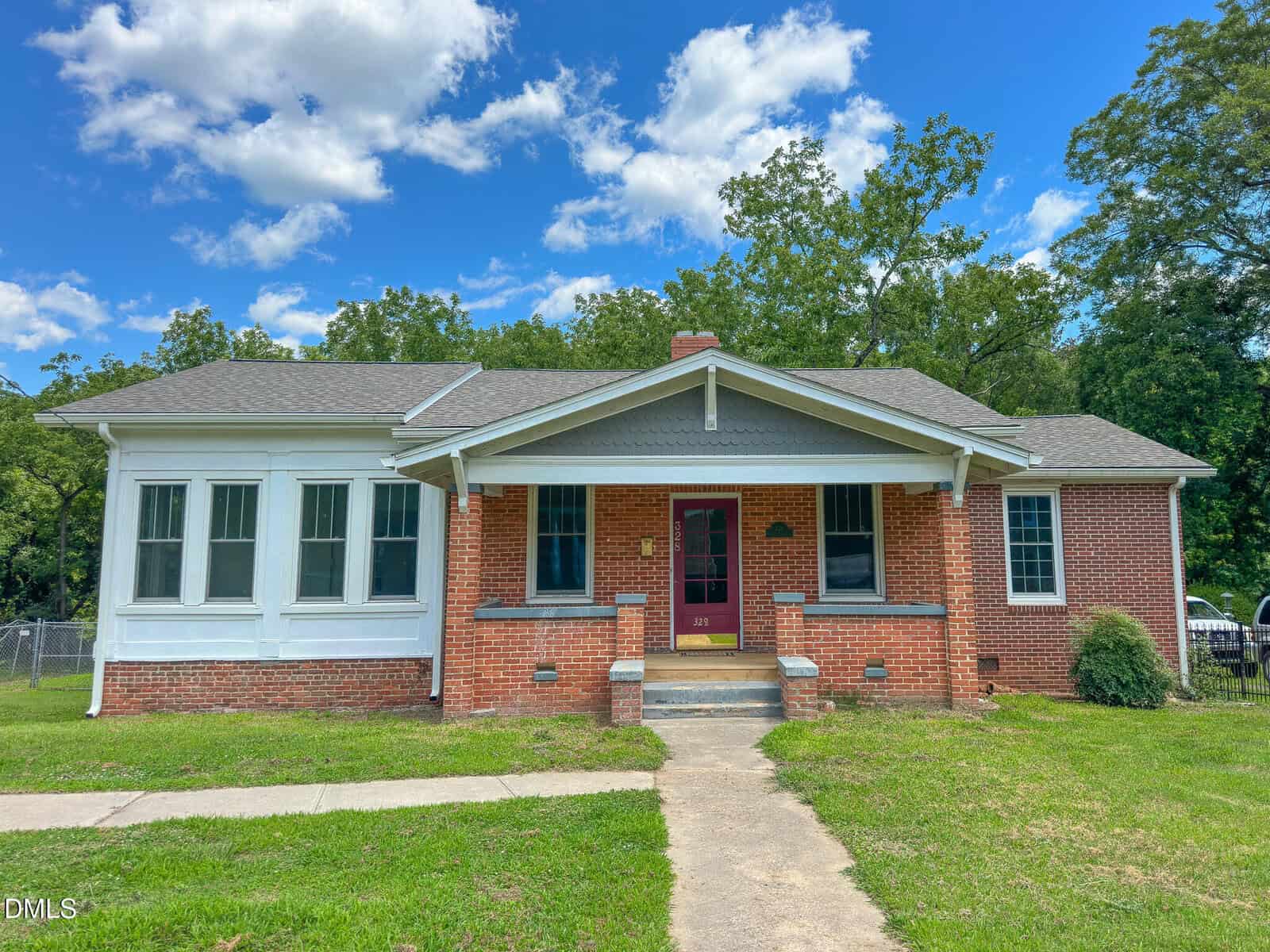 A red-brick house at 328 N Main Street in Warrenton features a covered front porch, white trim, a purple front door, and a grassy yard under a blue sky with scattered clouds. Trees and a fence are in the background.