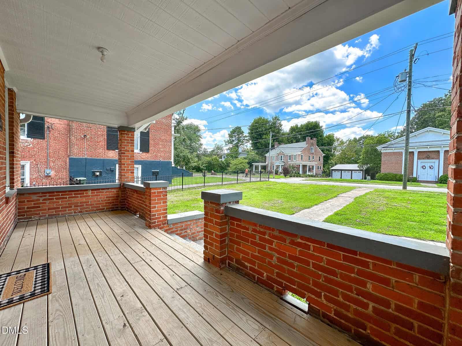 View from a covered porch with a wooden floor and brick railing, overlooking a green lawn, sidewalks, neighboring red-brick buildings, and a two-story house across the street under a partly cloudy sky.