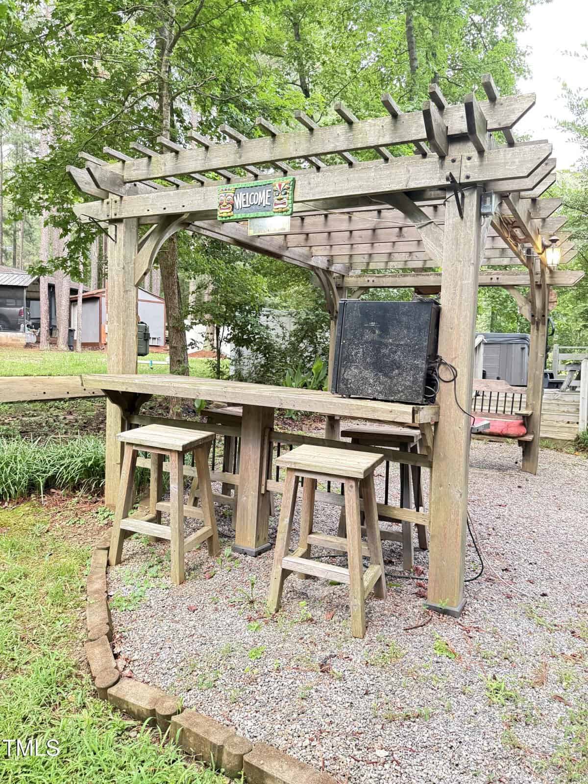 Wooden outdoor bar with three stools under a pergola, set on a gravel area. A “Welcome” sign hangs above, and a black mini fridge sits on the bar. Green trees and nearby houses are visible in the background.