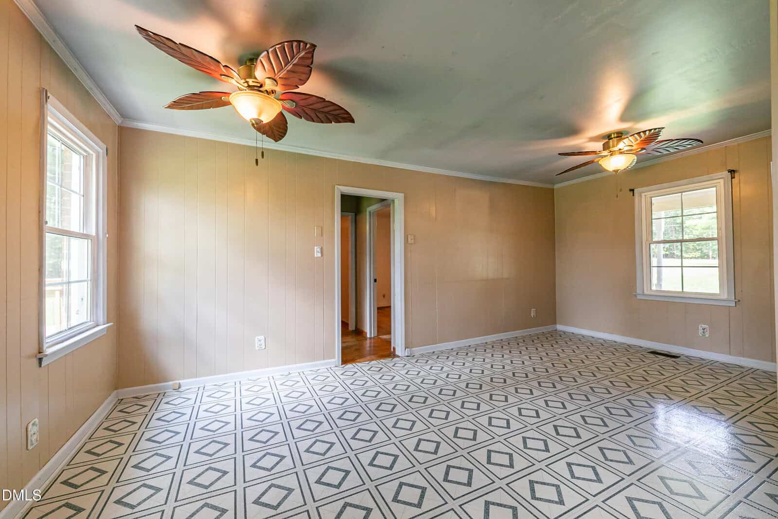 Empty room with beige paneled walls, patterned tile floor, two large windows, and two lighted ceiling fans with leaf-shaped blades. An open doorway leads to another room with wooden flooring.