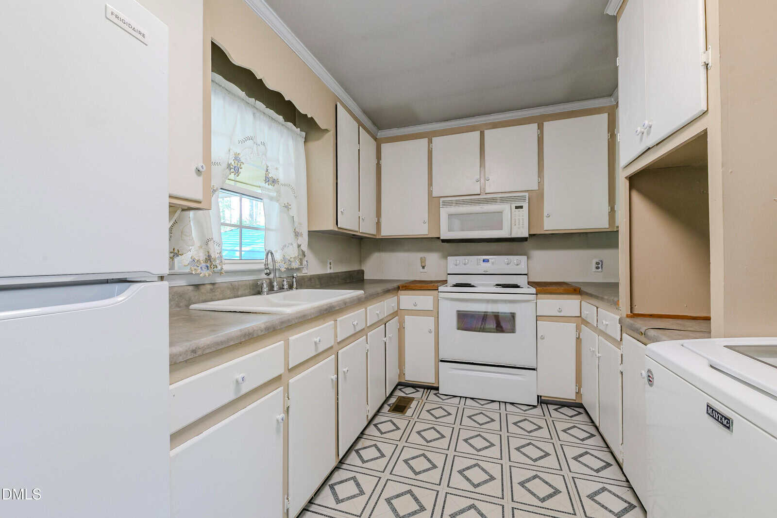A kitchen with white cabinets, beige countertops, a white stove with oven, a white microwave, white refrigerator, double sink under a window, and patterned vinyl flooring. Walls are light brown.