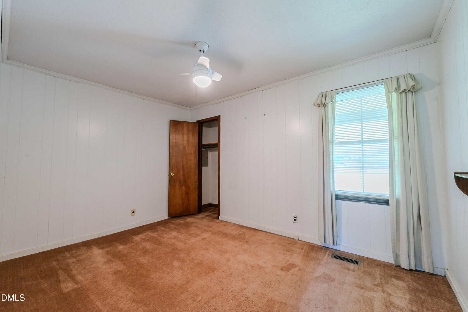 Empty room with tan carpet, white paneled walls, a ceiling fan, a window with white curtains, and a wooden door leading to a small closet. Natural light enters through the window.