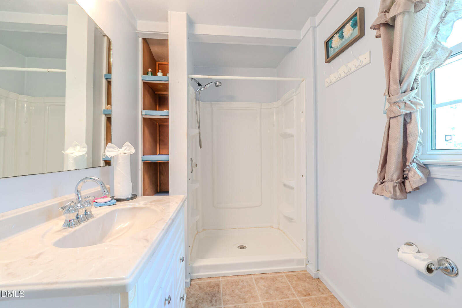 A bathroom with a white vanity and sink, an open shelving unit, a walk-in shower with a handheld showerhead, a window with curtains, and tiled flooring.