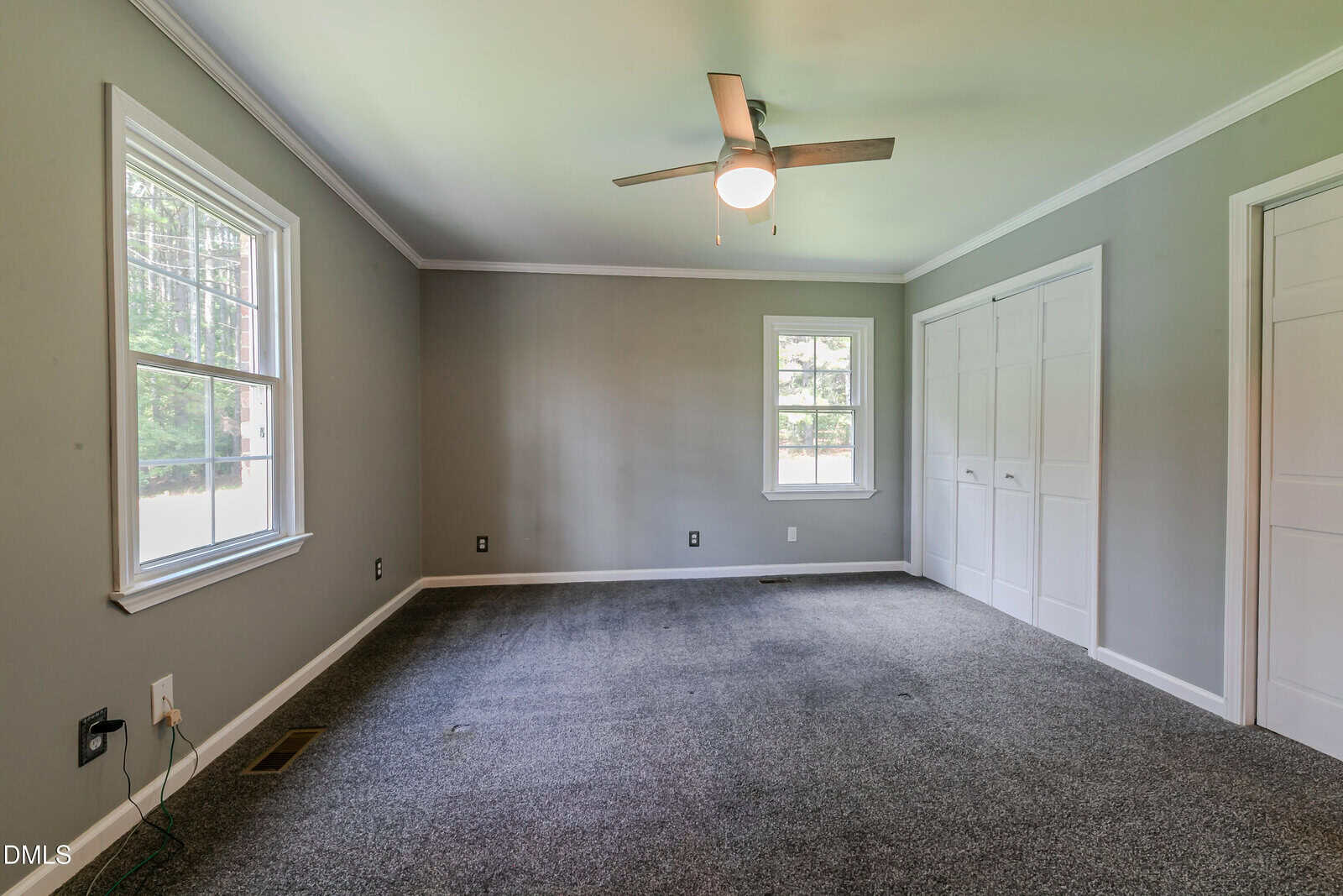 Empty bedroom with gray walls, gray carpet, a ceiling fan with light, two windows, white trim, and closed white double closet doors. There are visible electrical outlets and natural light from the windows.