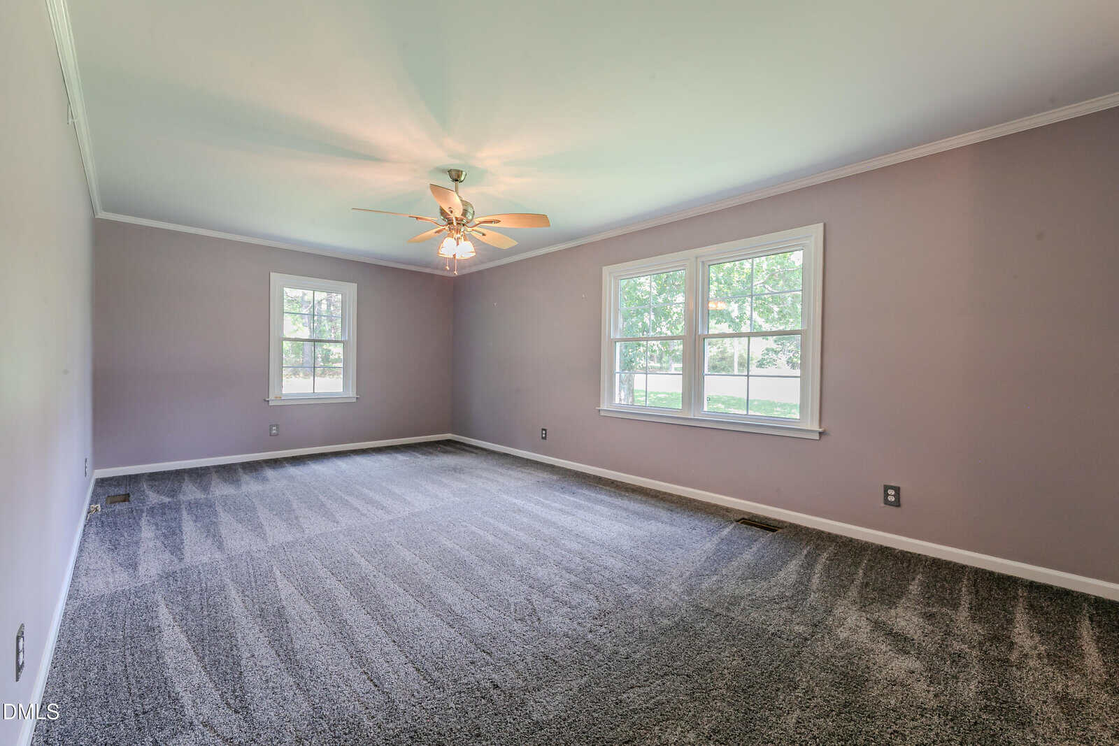 Empty room with light purple walls, gray carpet, two windows on one side, one window on another, and a ceiling fan with lights. The room is well-lit by natural light and has white trim.