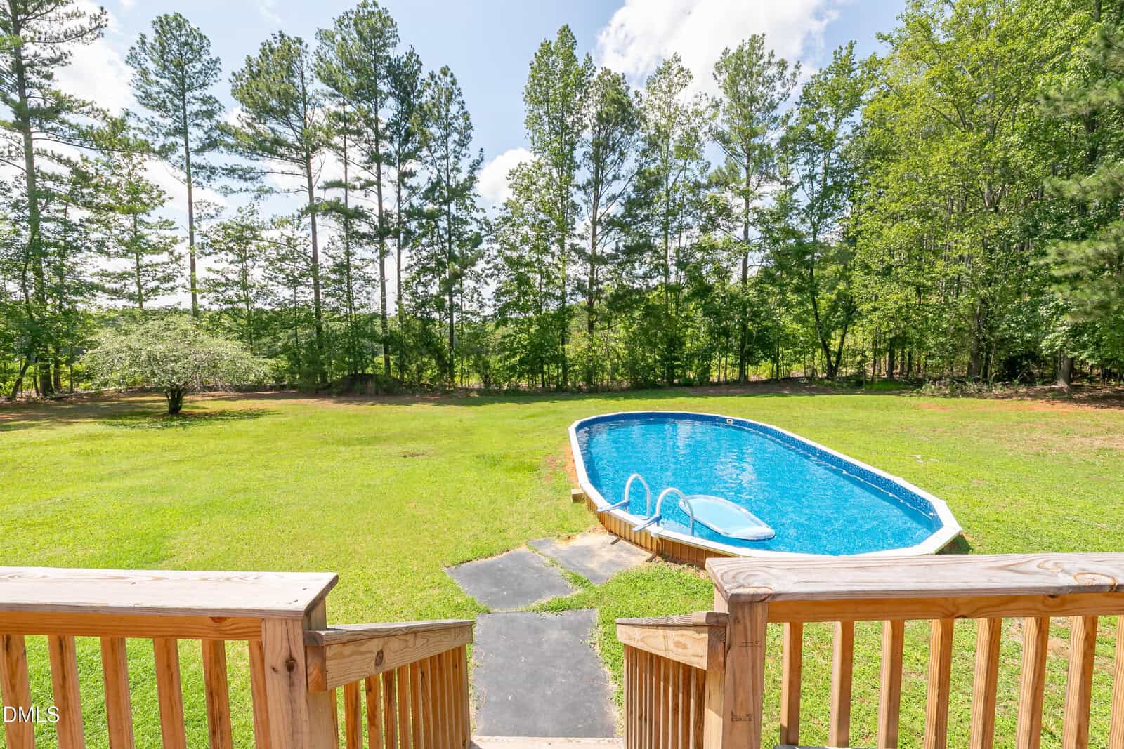 A wooden deck overlooks a large, grassy backyard with an above-ground oval pool. Tall trees border the yard, and the sky is partly cloudy. Steps lead down from the deck to the pool area.