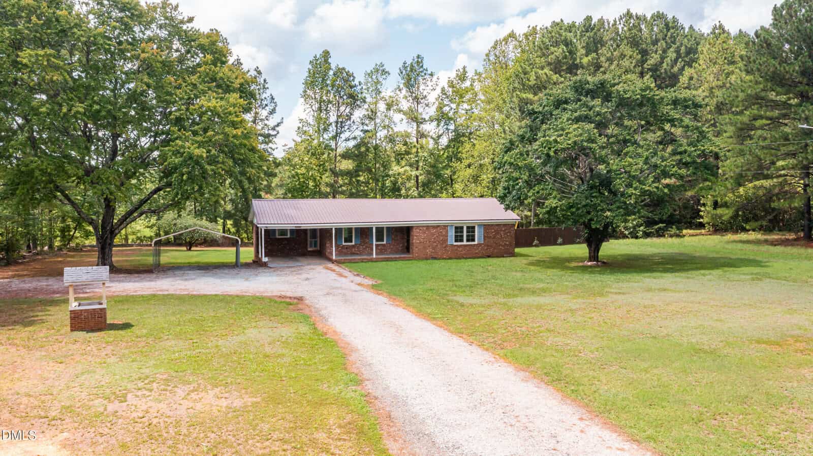 Single-story brick house with a metal roof, surrounded by grass and trees at 1751 Mountain Creek Road in Oxford. A gravel driveway leads to the house, with a mailbox on the left and a carport near the entrance. The setting appears rural and spacious.