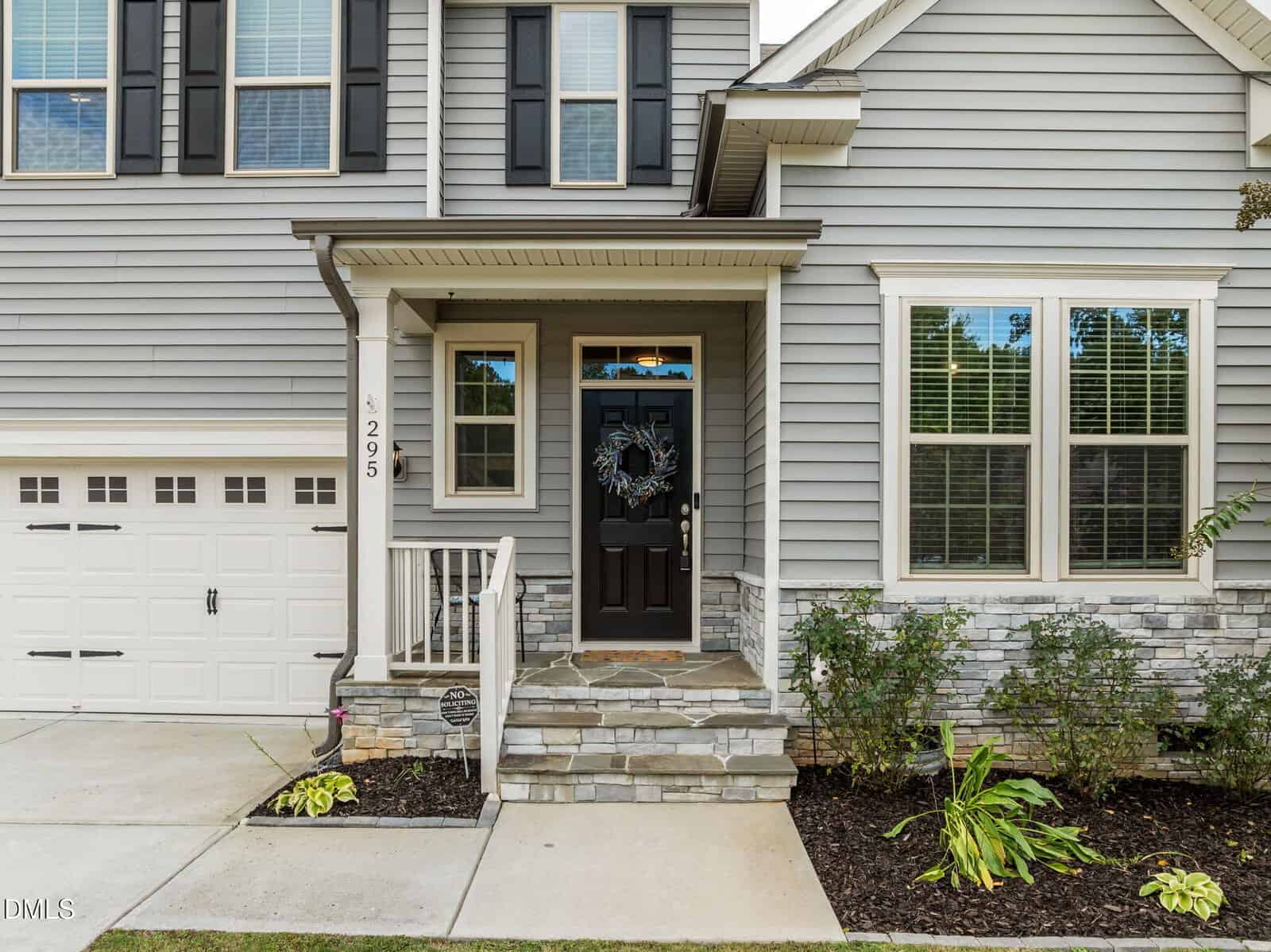 Front view of a gray two-story house with black shutters, a black front door with a wreath, stone steps, a small porch, attached garage, and landscaped plants along the walkway.