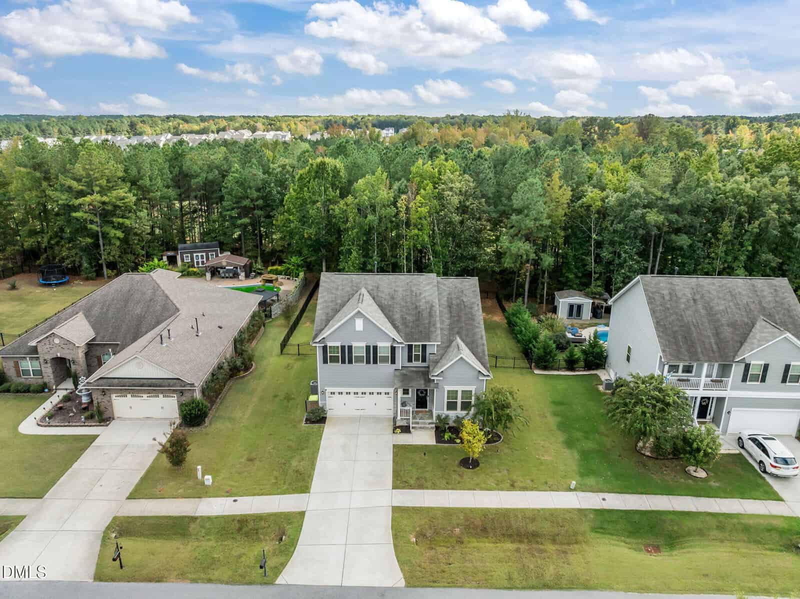 Aerial view of three suburban houses with driveways, lawns, and landscaping, bordered by a wooded area in the background under a partly cloudy sky.