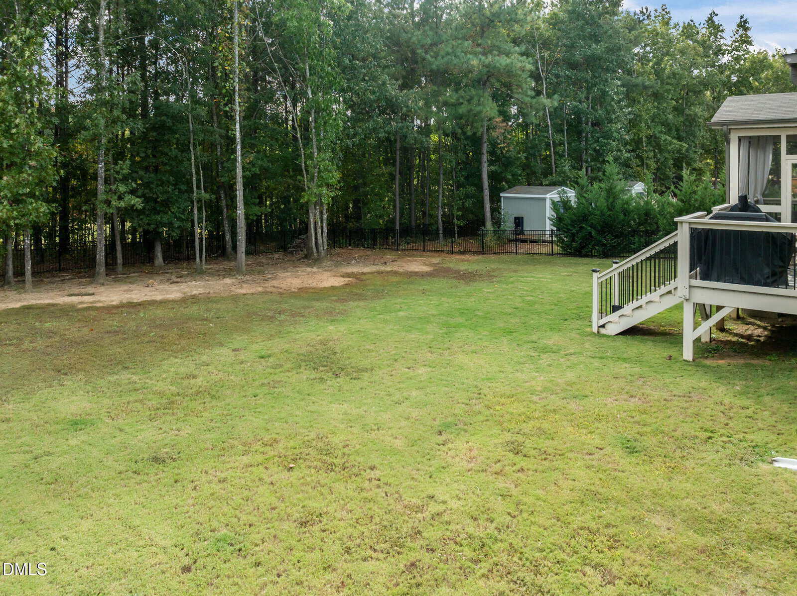 A backyard with a grassy lawn, a small storage shed near trees, and a white-railed staircase leading from a deck attached to a house. Forested area borders the yard in the background.
