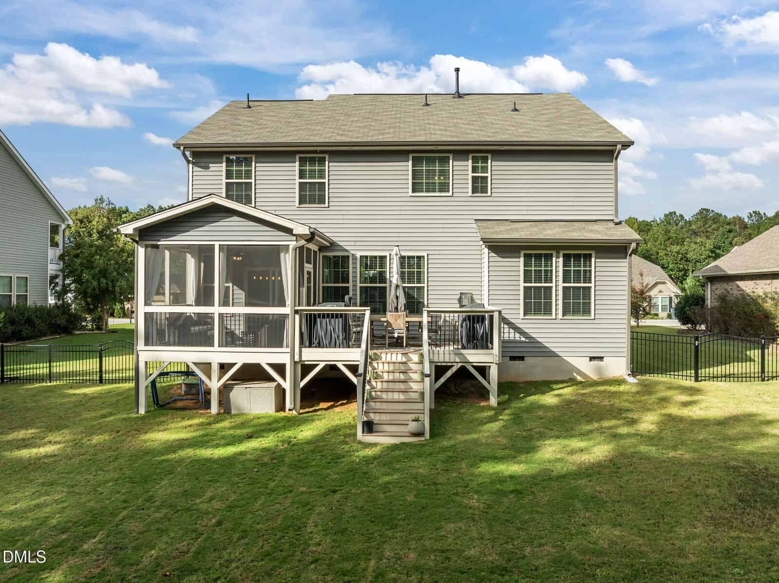 Two-story gray house with white trim, screened porch, open deck, stairs leading to a grassy backyard, and black metal fence, under a blue sky with scattered clouds.